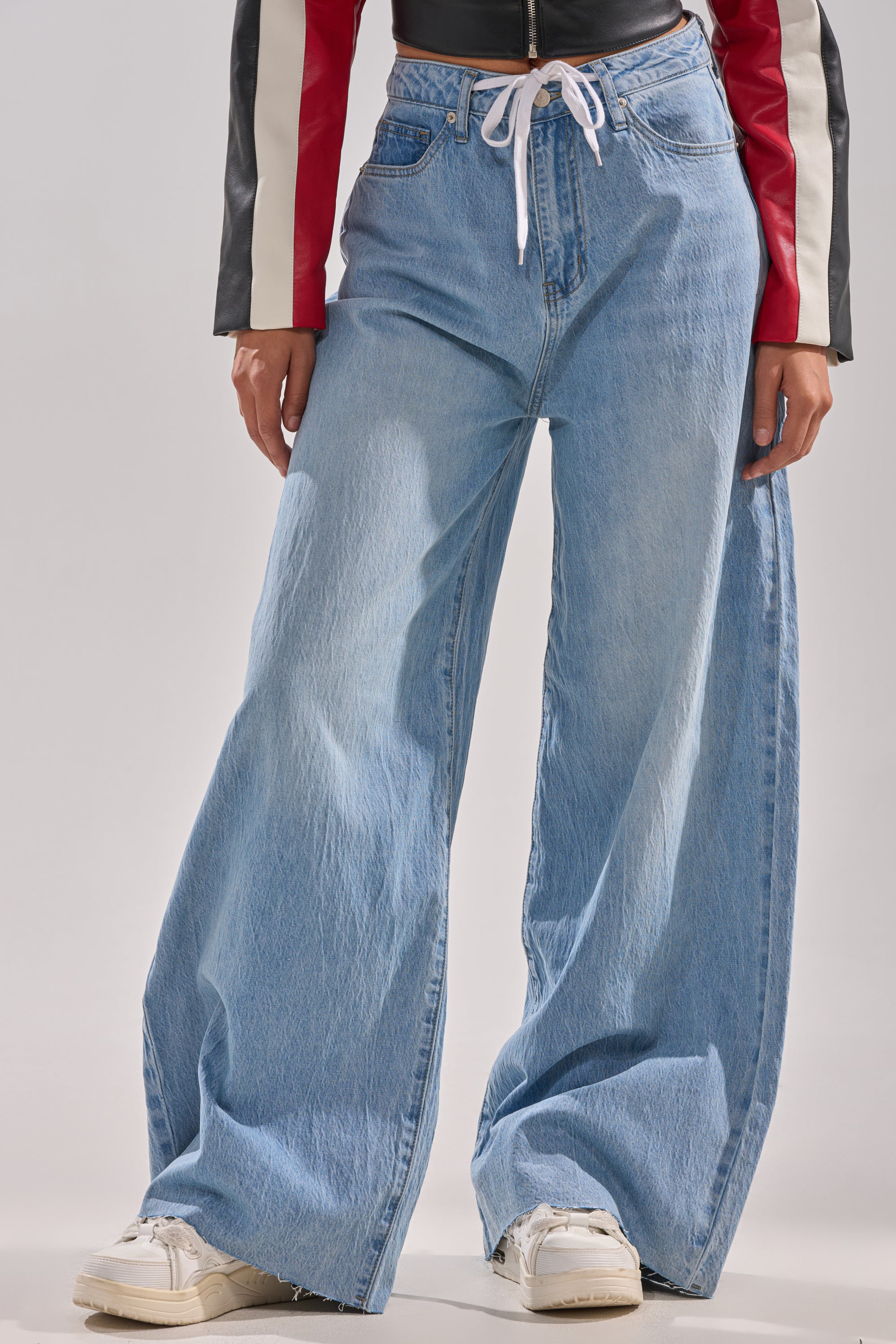 Wearing YOU BET DRAWSTRING WIDE LEG JEANS in light blue, paired with a cropped jacket with black, white, and red sleeves and white sneakers. The setting features a plain light background.