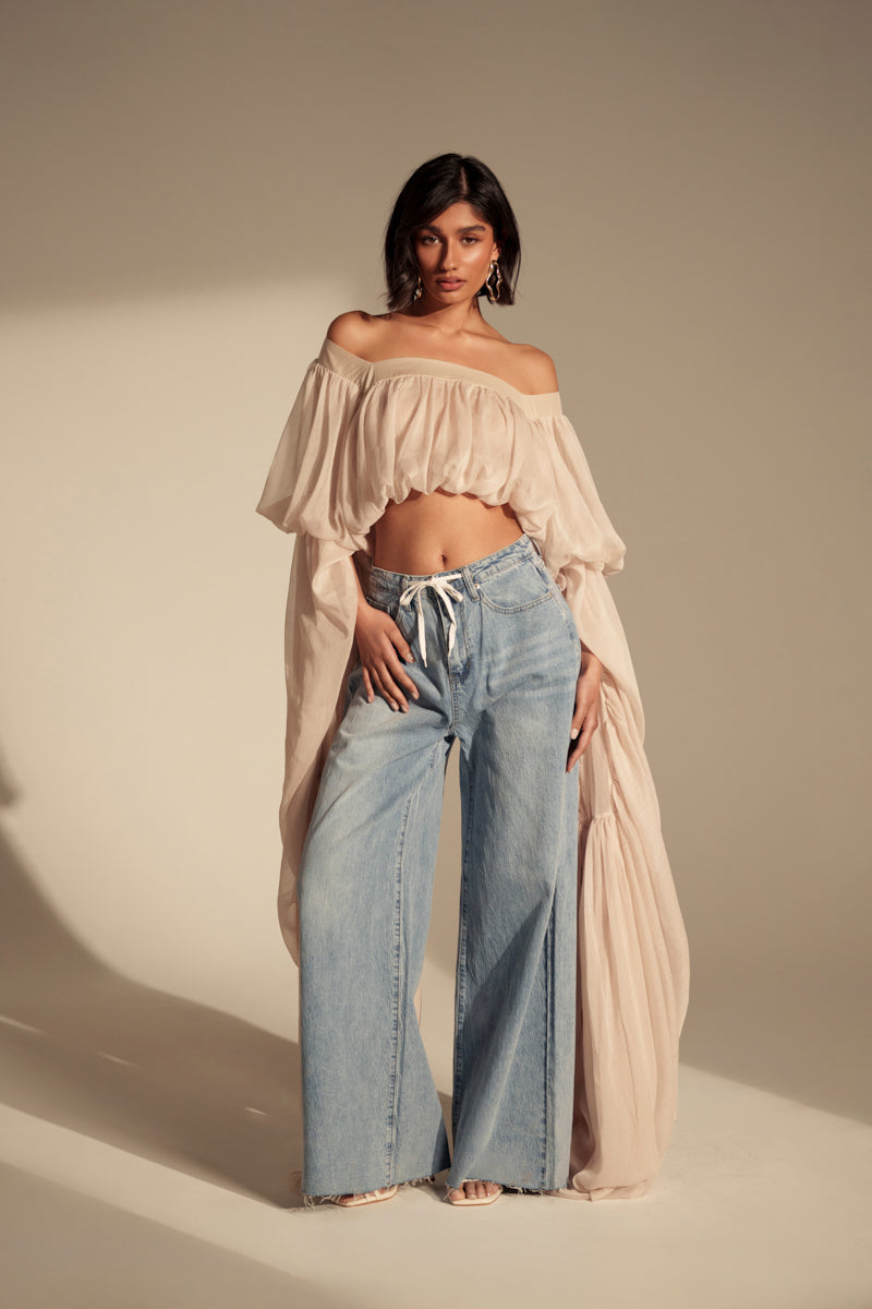A woman poses against a plain beige background in vintage-wash YOU BET DRAWSTRING WIDE LEG JEANS, styled with a loose off-the-shoulder cream crop top, long billowy sleeves, hoop earrings, and heels.