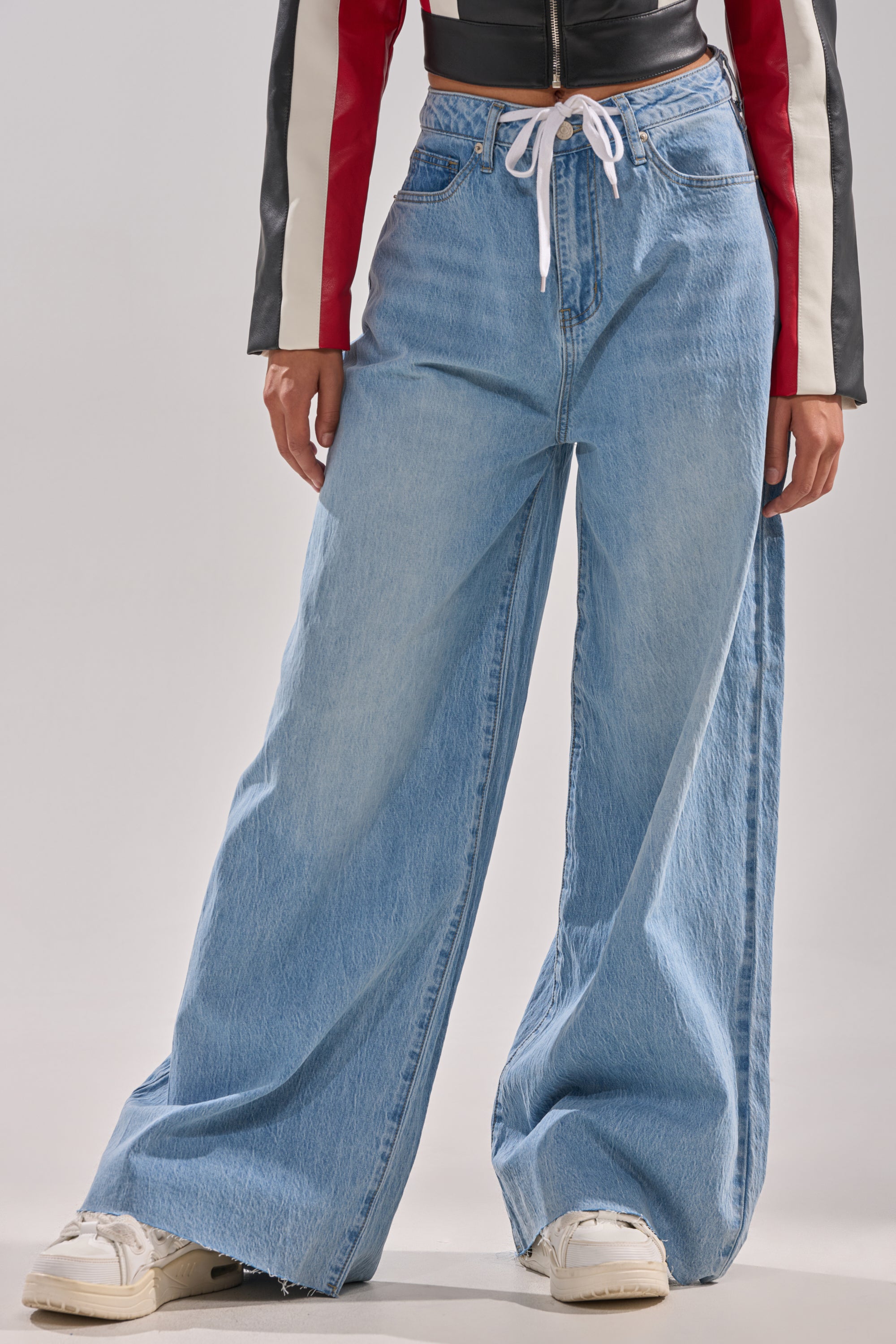 Someone wearing YOU BET DRAWSTRING WIDE LEG JEANS—vintage wash denim with a white drawstring waist and frayed hems—paired with white sneakers and a red, black, and white long-sleeve jacket.