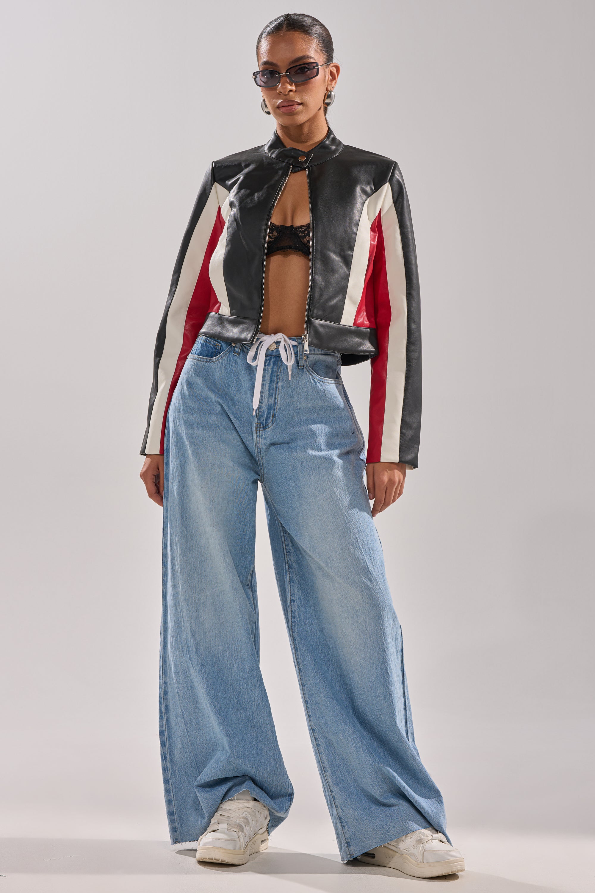 A woman stands facing forward against a plain background, wearing a black, red, and white leather jacket over a black bralette, YOU BET DRAWSTRING WIDE LEG JEANS, white sneakers, and sunglasses.