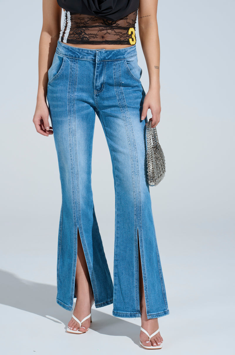 Wearing SPLIT HEM BOOTCUT TROUSER JEAN, a black lace top, white heeled sandals, and holding a small silver handbag, a person stands against a plain light background.