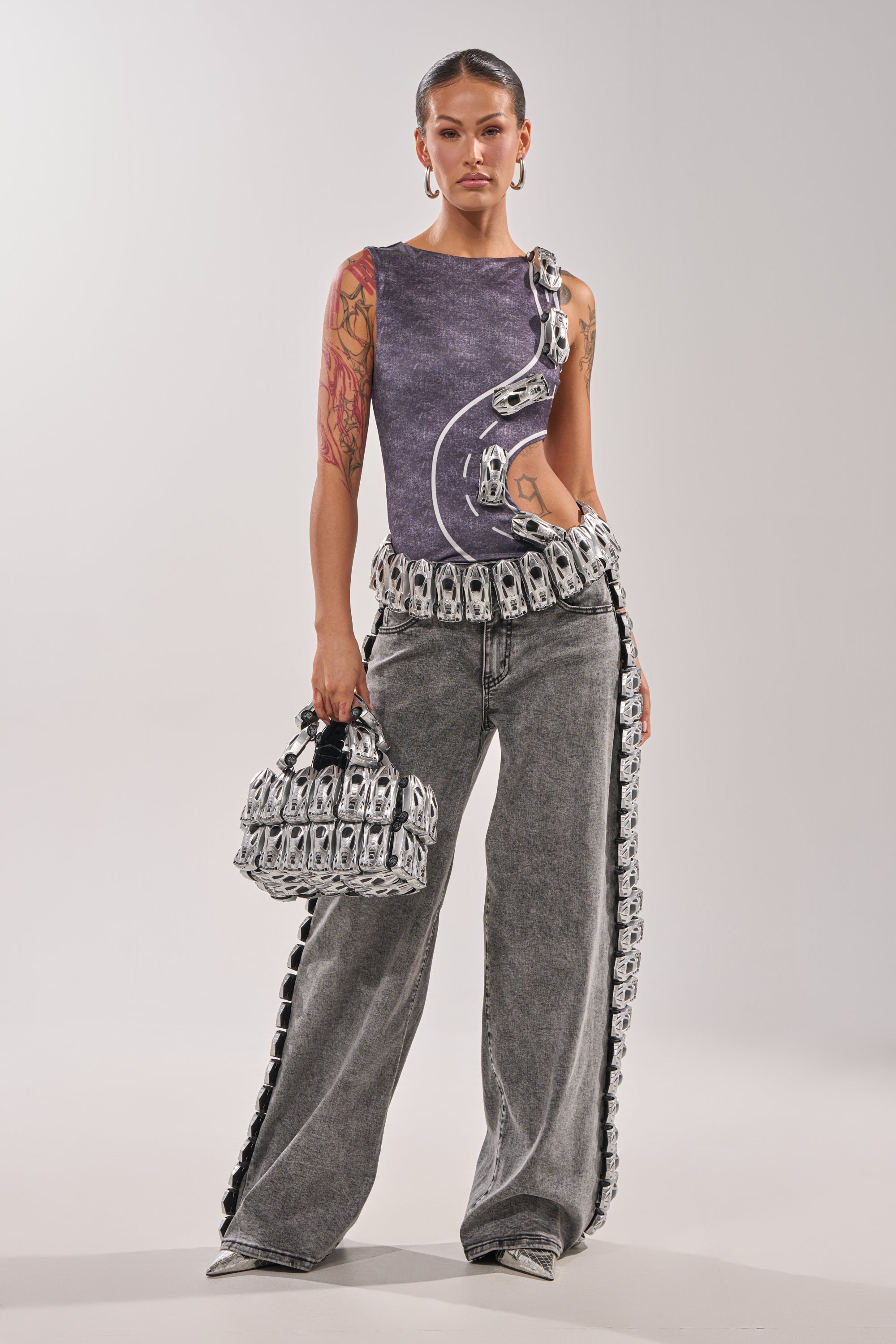 A woman with slicked-back hair and hoop earrings stands forward, wearing a sleeveless purple cut-out top, wide leg RACE CAR DENIM jeans with metallic detailing, and holding a matching handbag. 1