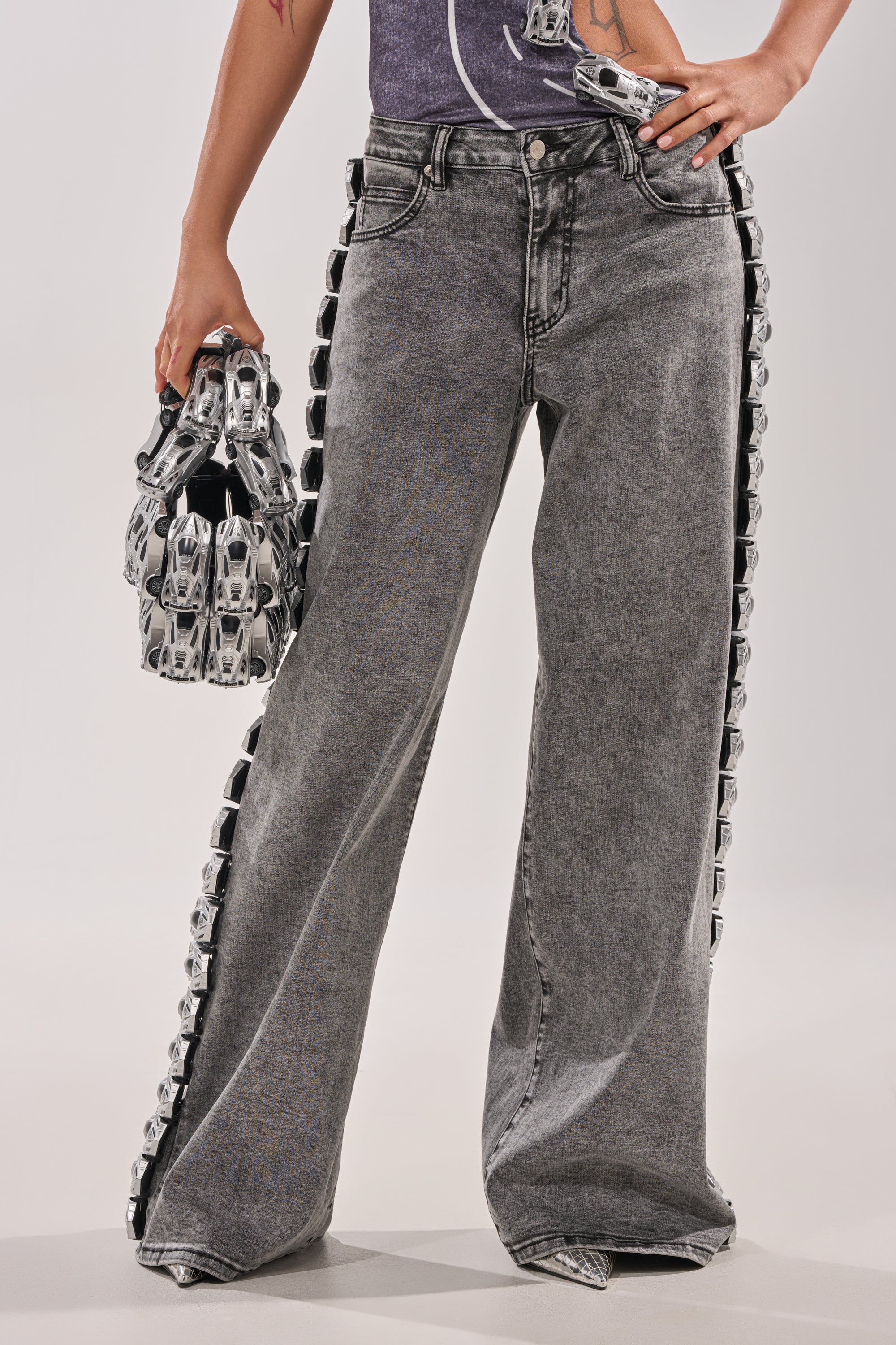 A person wears RACE CAR DENIM—wide leg, race car–inspired jeans with shiny silver toy cars down each side—and holds a matching handbag made of toy cars. 3