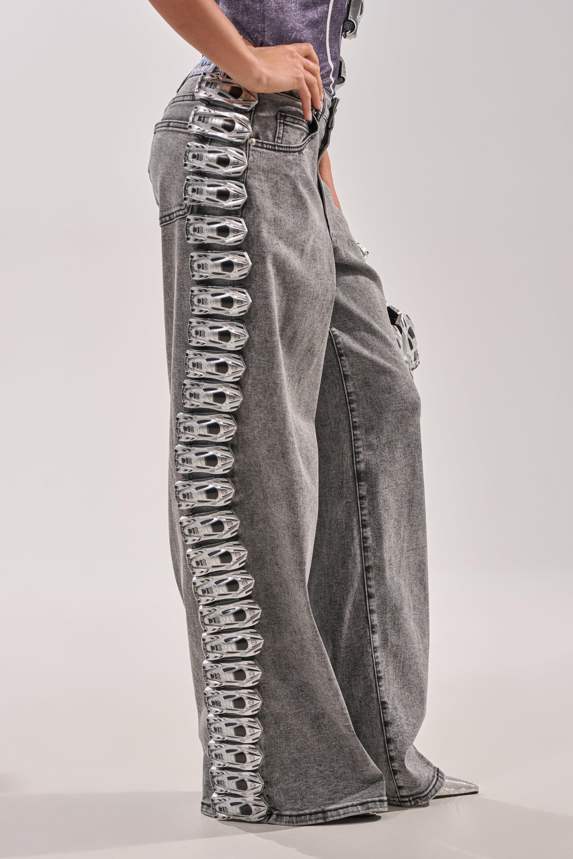 Wearing RACE CAR DENIM—wide-leg gray jeans featuring a row of metallic can pull tabs along the outer seams—a person stands against a plain light background. 0