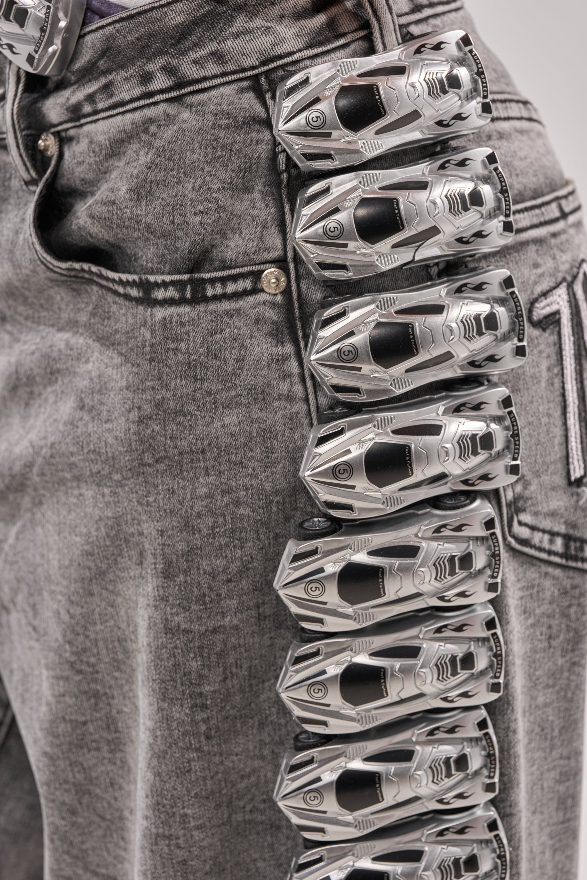 Close-up of RACE CAR DENIM styled with a belt made of linked silver toy cars, giving wide leg jeans a playful streetwear vibe. 7