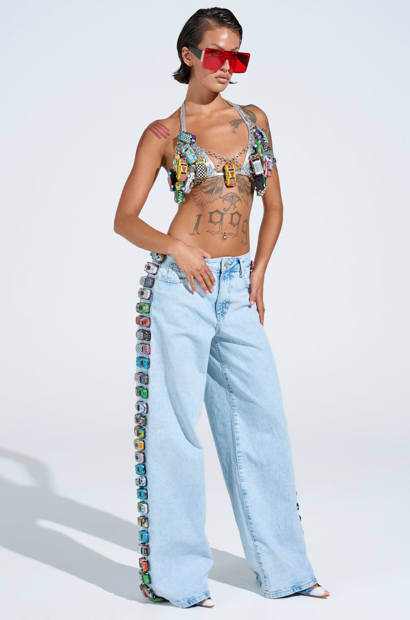 A person confidently wears red sunglasses, a colorful embellished bralette, and RACE CAR JEANS featuring small toy cars on the sides. They stand against a plain background, rocking their jeans with flair.