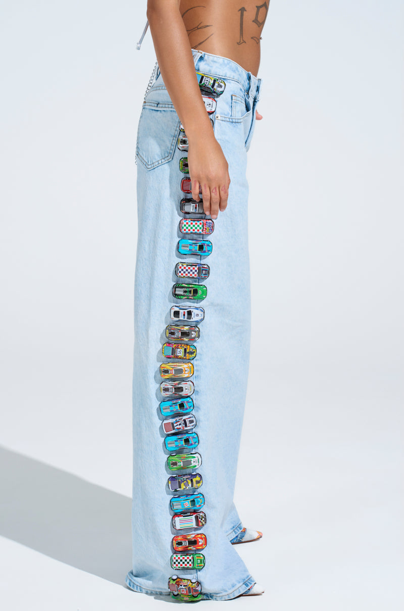 A person stands in relaxed wide-leg, light wash RACE CAR JEANS, adorned with a playful row of colorful toy cars along the outer seam of the right leg. The background is plain light blue, enhancing the jeans' charm. The upper body is partially visible.