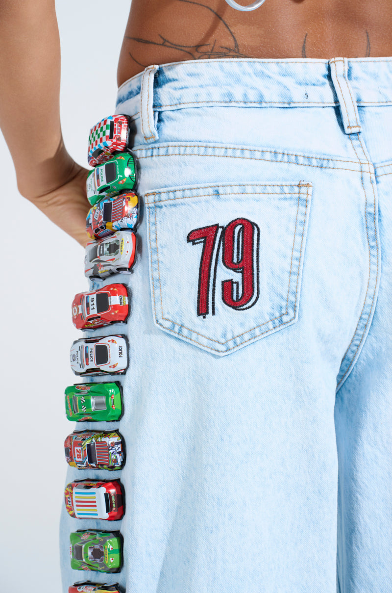A person wears RACE CAR JEANS, featuring relaxed wide-leg light wash denim with "79" embroidered on the back pocket and a vertical line of toy race cars from waist to knee, set against a plain white background.