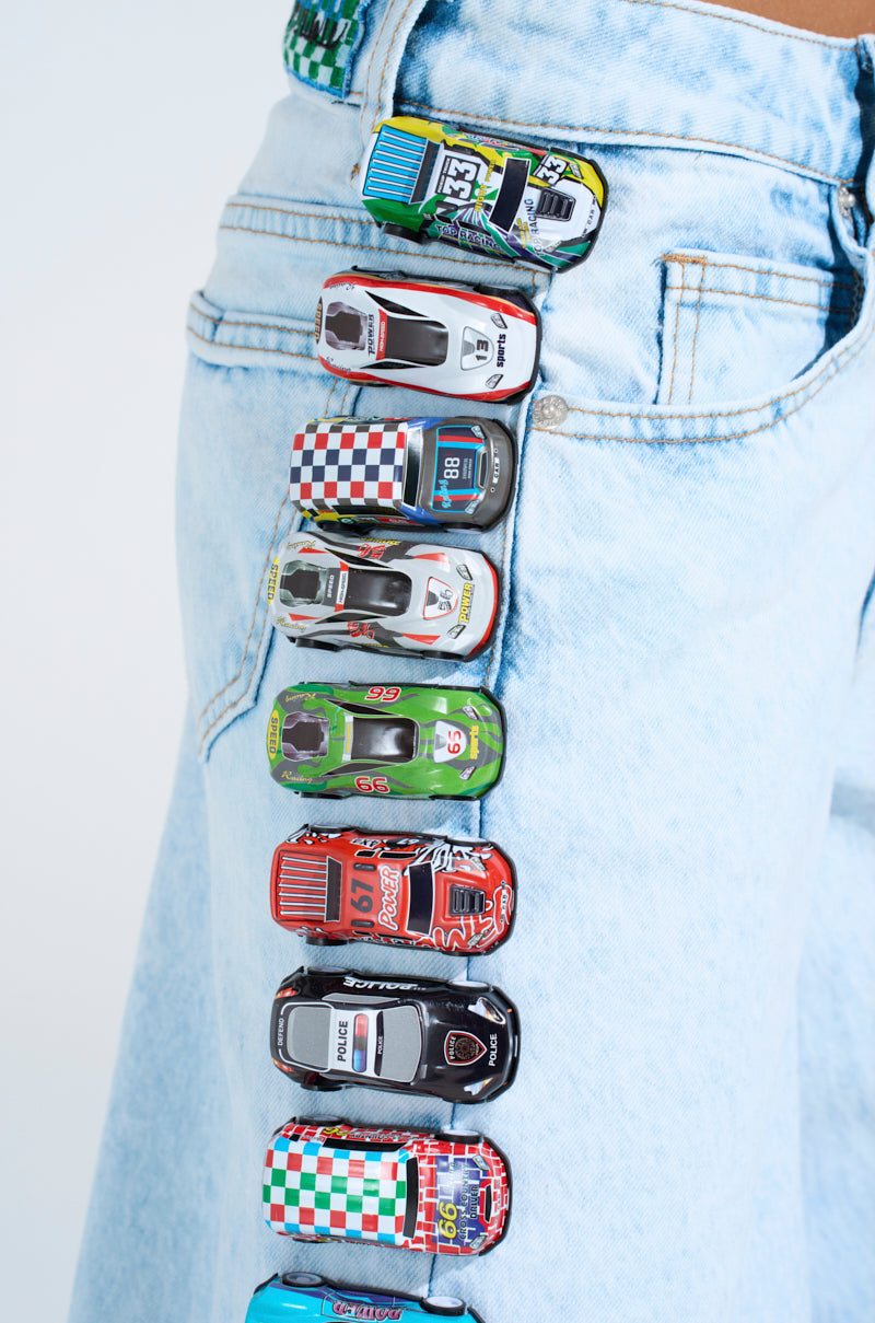 A row of vibrant toy race cars lines the fold of relaxed wide-leg jeans, the light wash denim showcasing playful designs like checkered patterns and numbers. These are the RACE CAR JEANS in full display.