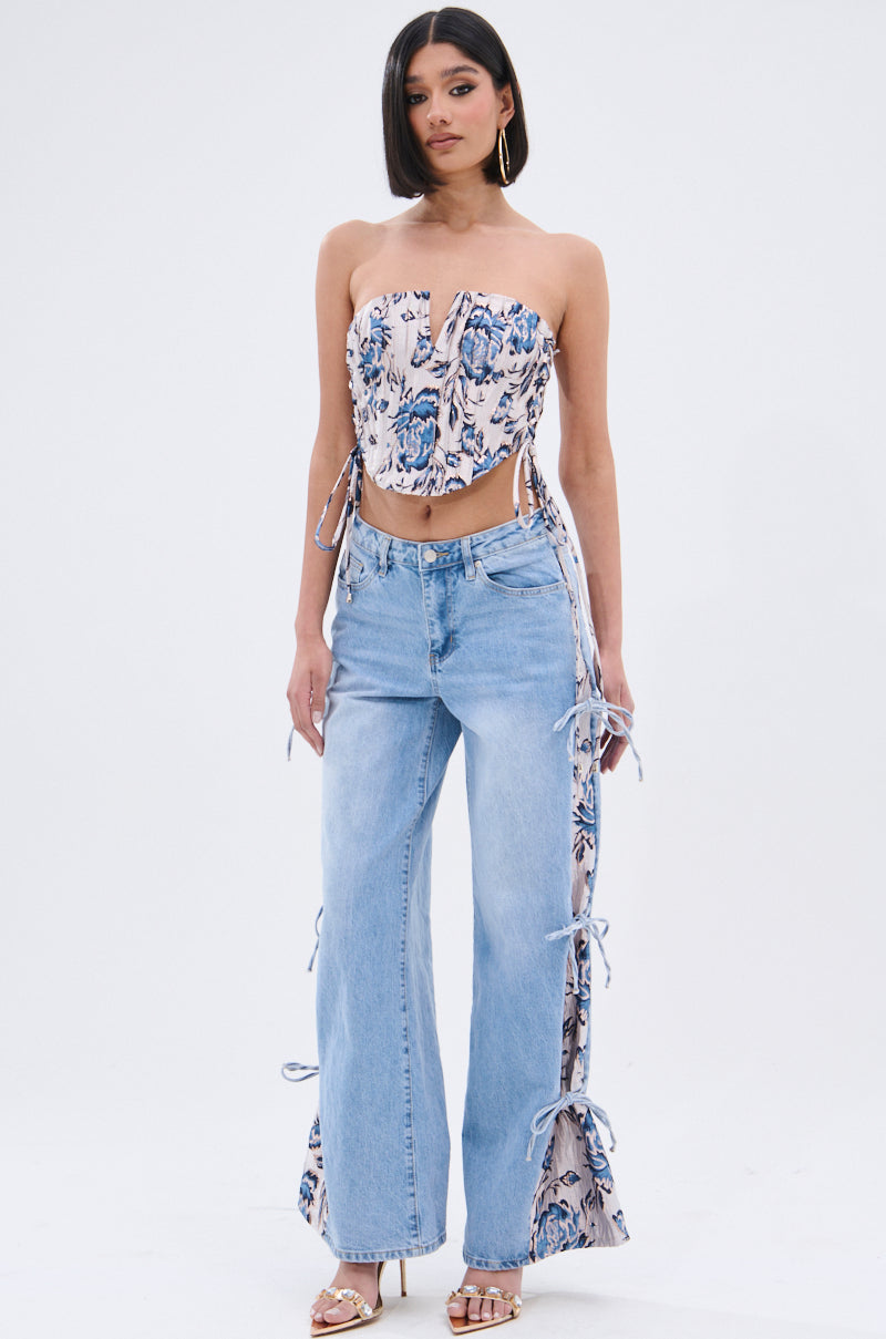 A woman poses against a white background in a strapless floral corset top and SEASONAL AWAKENING BROCADE PATCH DENIM JEANS featuring side slits tied with bows. She finishes her look with open-toe heels and gold hoop earrings. 0