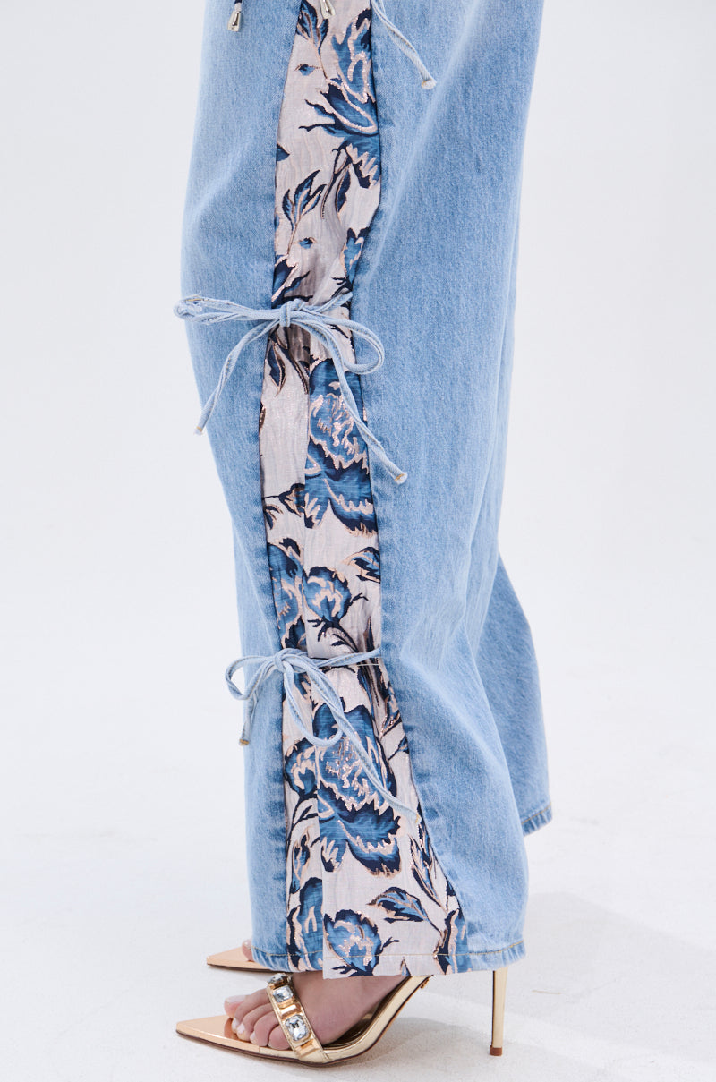 Someone is wearing SEASONAL AWAKENING BROCADE PATCH DENIM JEANS—light blue high-rise wide-leg jeans with a floral brocade panel and side seam bows—paired with open-toe heeled sandals featuring a decorative strap. 9
