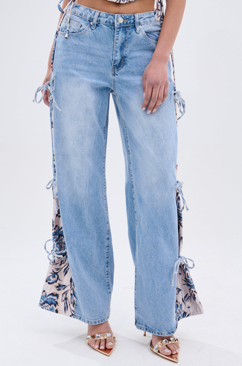 A person wears SEASONAL AWAKENING BROCADE PATCH DENIM JEANS—light blue, high-rise wide-leg jeans with side cutouts tied by bows that reveal floral brocade denim. They pair them with open-toe heels with gold accents against a plain white background. 2