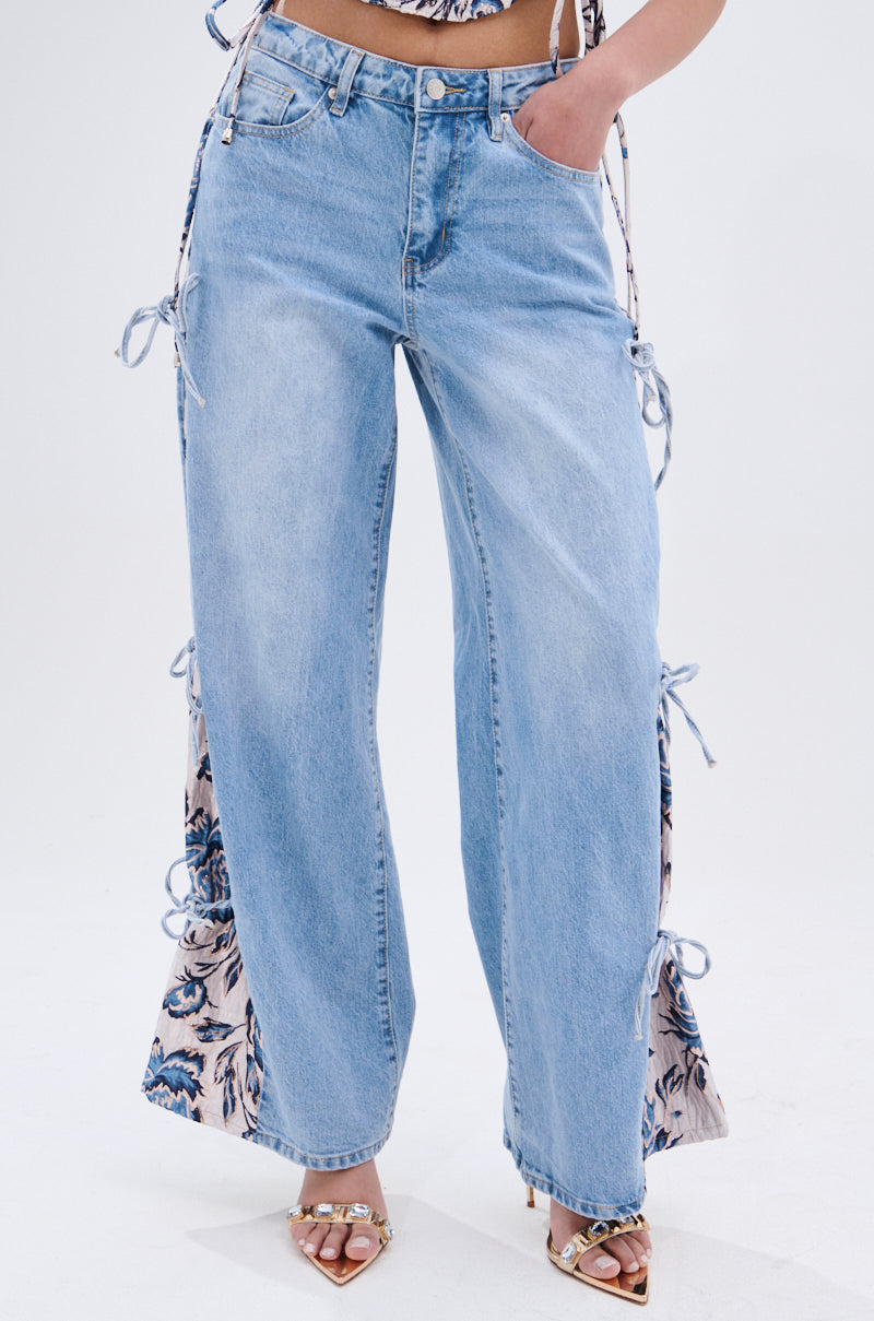 Wearing SEASONAL AWAKENING BROCADE PATCH DENIM JEANS—high-rise wide-leg jeans with light blue floral brocade panels and bow tie details on the sides—paired with heeled sandals; upper body is mostly out of frame. 3