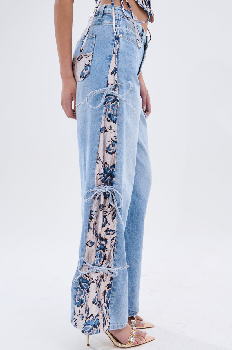 A person wears SEASONAL AWAKENING BROCADE PATCH DENIM JEANS with high-rise wide legs, floral brocade panels tied by white laces, a floral pocket, and strappy heels featuring gold chain details. 5