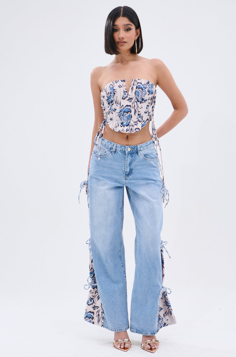 A woman poses against a white background in a floral strapless corset top paired with SEASONAL AWAKENING BROCADE PATCH DENIM JEANS—light blue, high-rise, wide-leg jeans with lace-up sides—styled with heels, hoops, and straight shoulder-length hair. 7