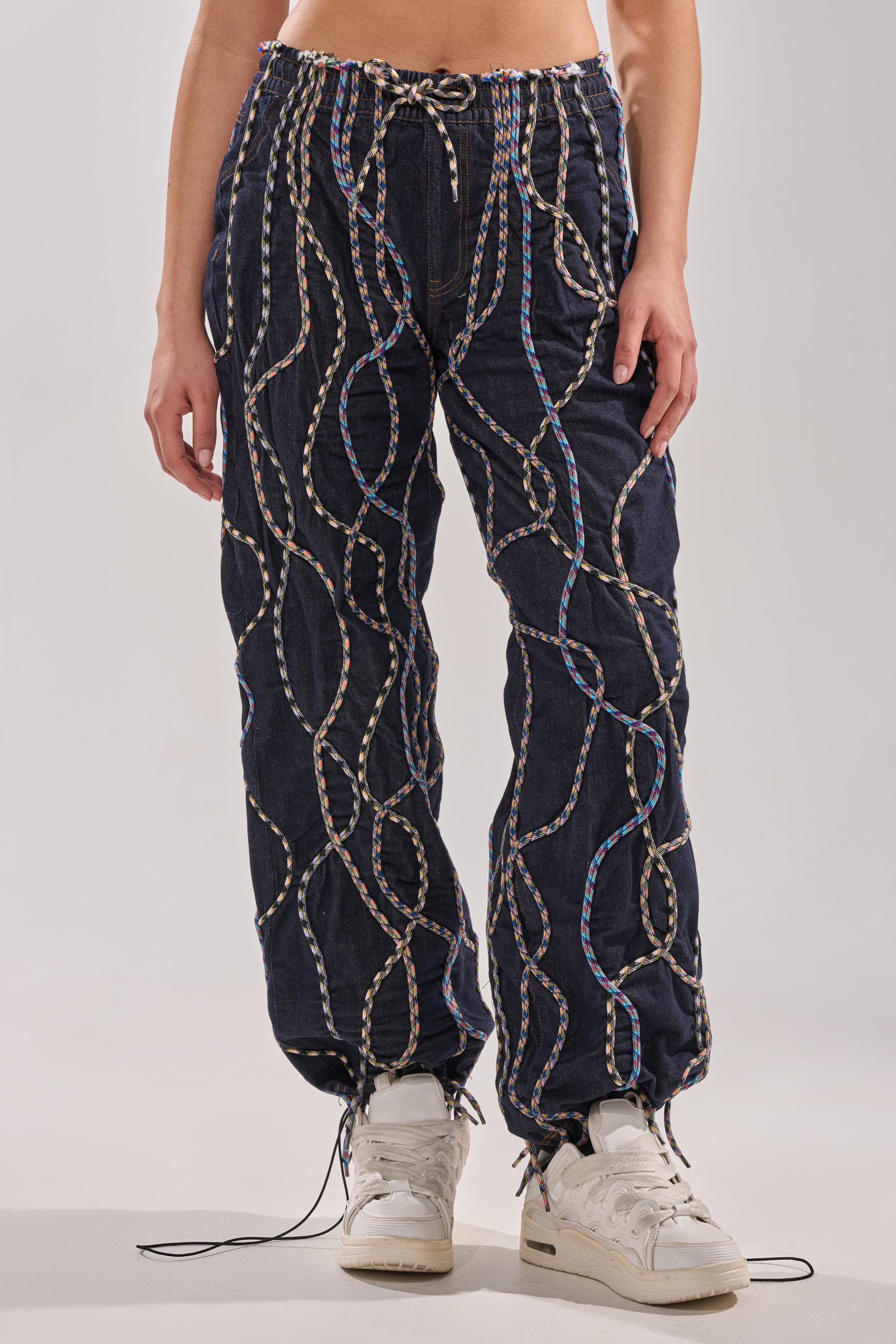 A person wears the CRAZY FOR YOU DENIM PANTS—navy blue, high-rise, loose-fit with multicolor rope detailing and drawstrings at the waist and ankles—paired with white sneakers against a plain background. 0
