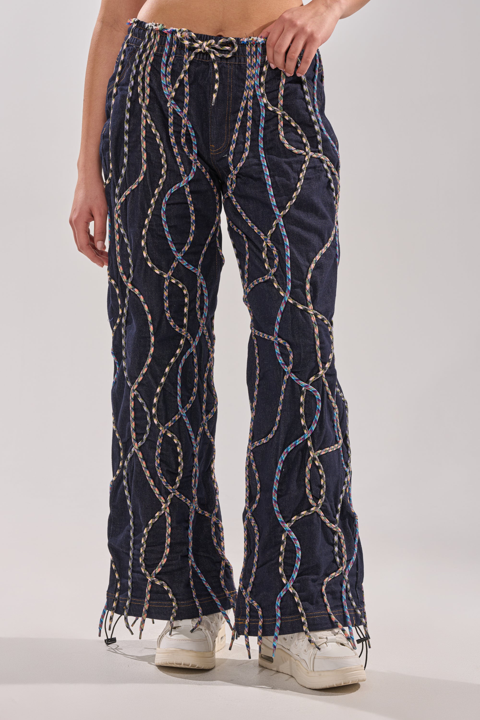 A person wears CRAZY FOR YOU DENIM PANTS—high-rise, dark blue denim with multicolor rope details—paired with white sneakers. The photo is cropped at the torso and ankles, emphasizing the pants’ unique embellished design. 1