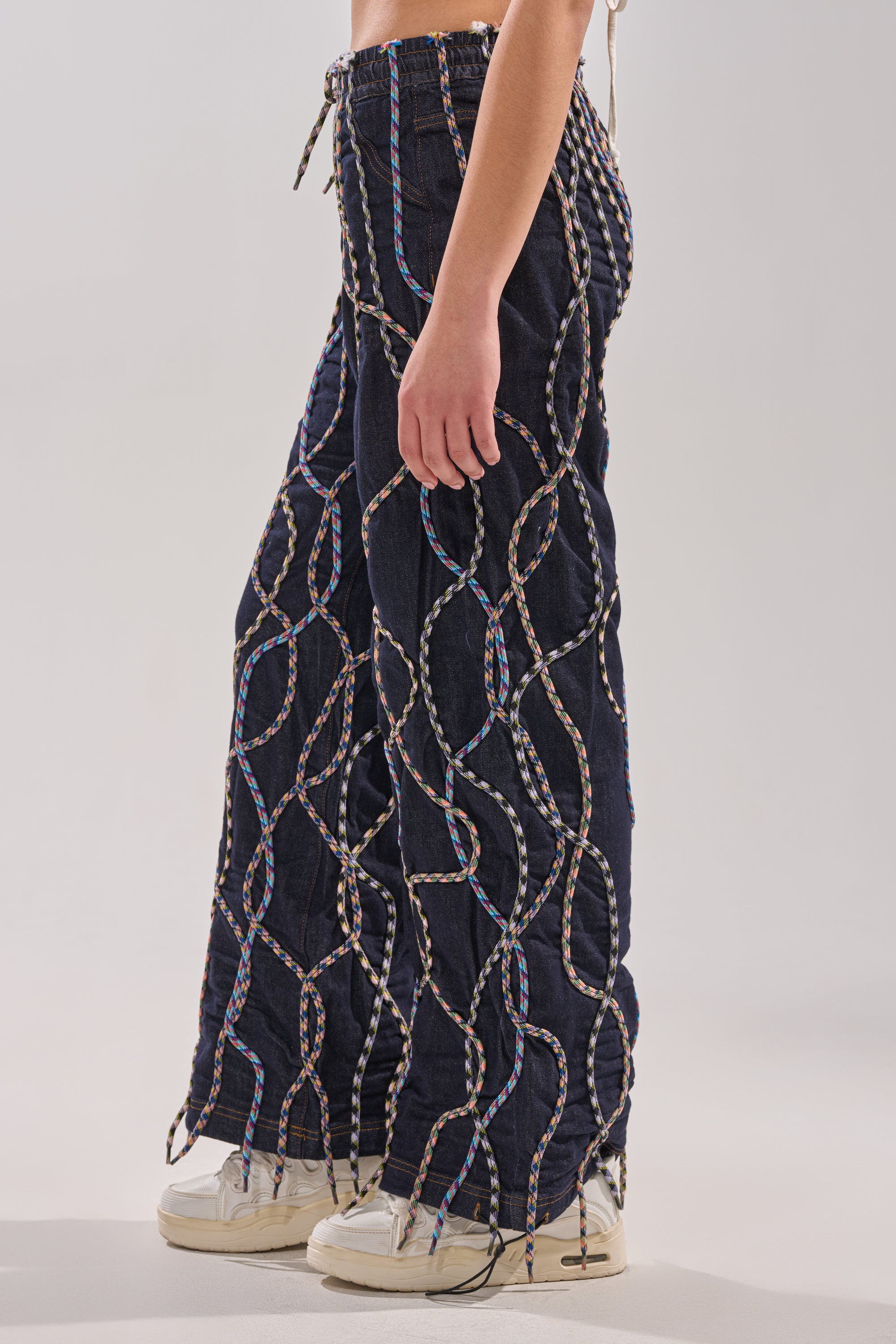 A person wearing the CRAZY FOR YOU DENIM PANTS—high-rise wide-leg navy blue pants with multicolor rope accents—paired with white sneakers, shown in a side view against a plain background. 2