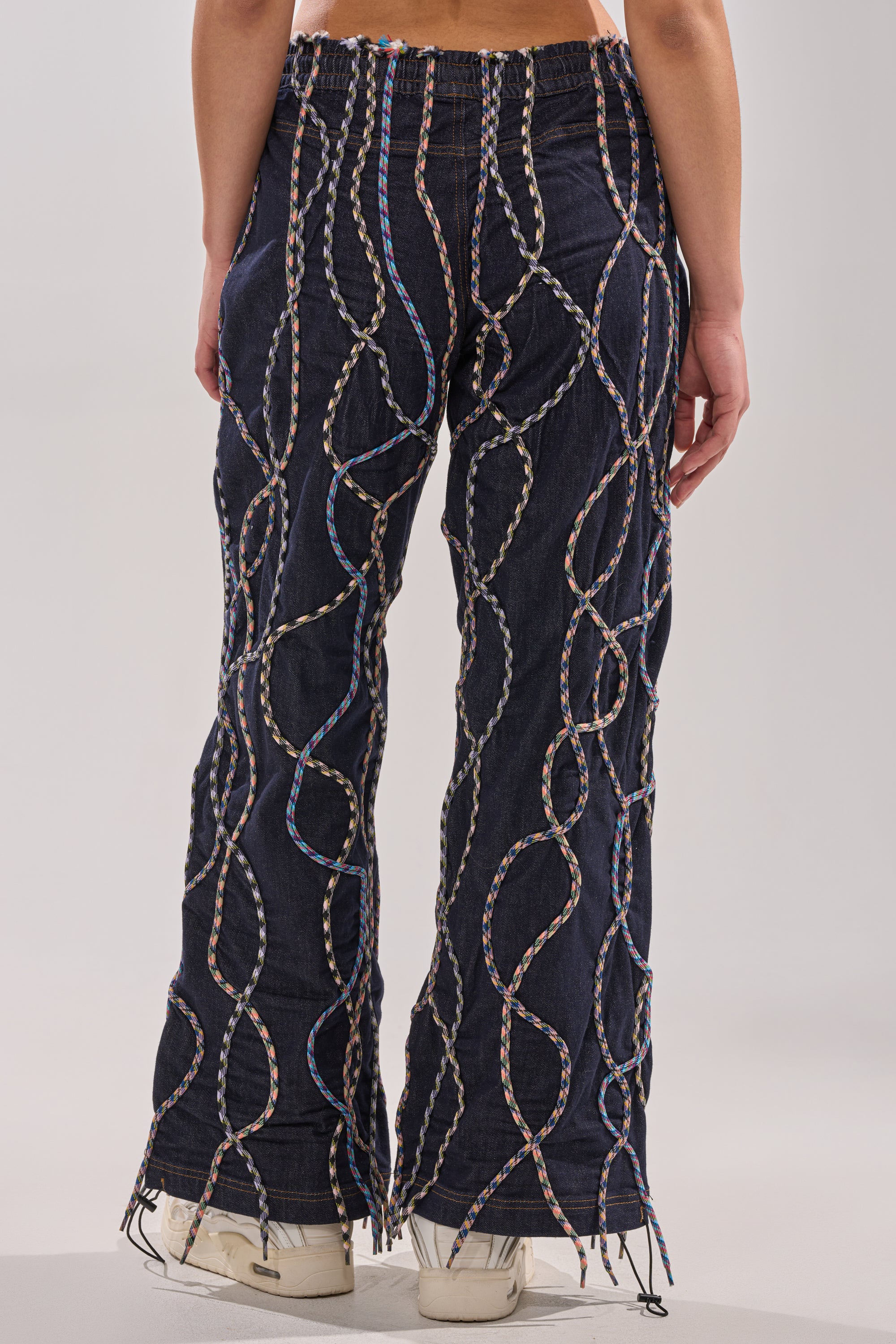 A person seen from the back wears CRAZY FOR YOU DENIM PANTS, featuring high-rise denim with multicolor rope stitched in looping patterns, paired with white sneakers. 3