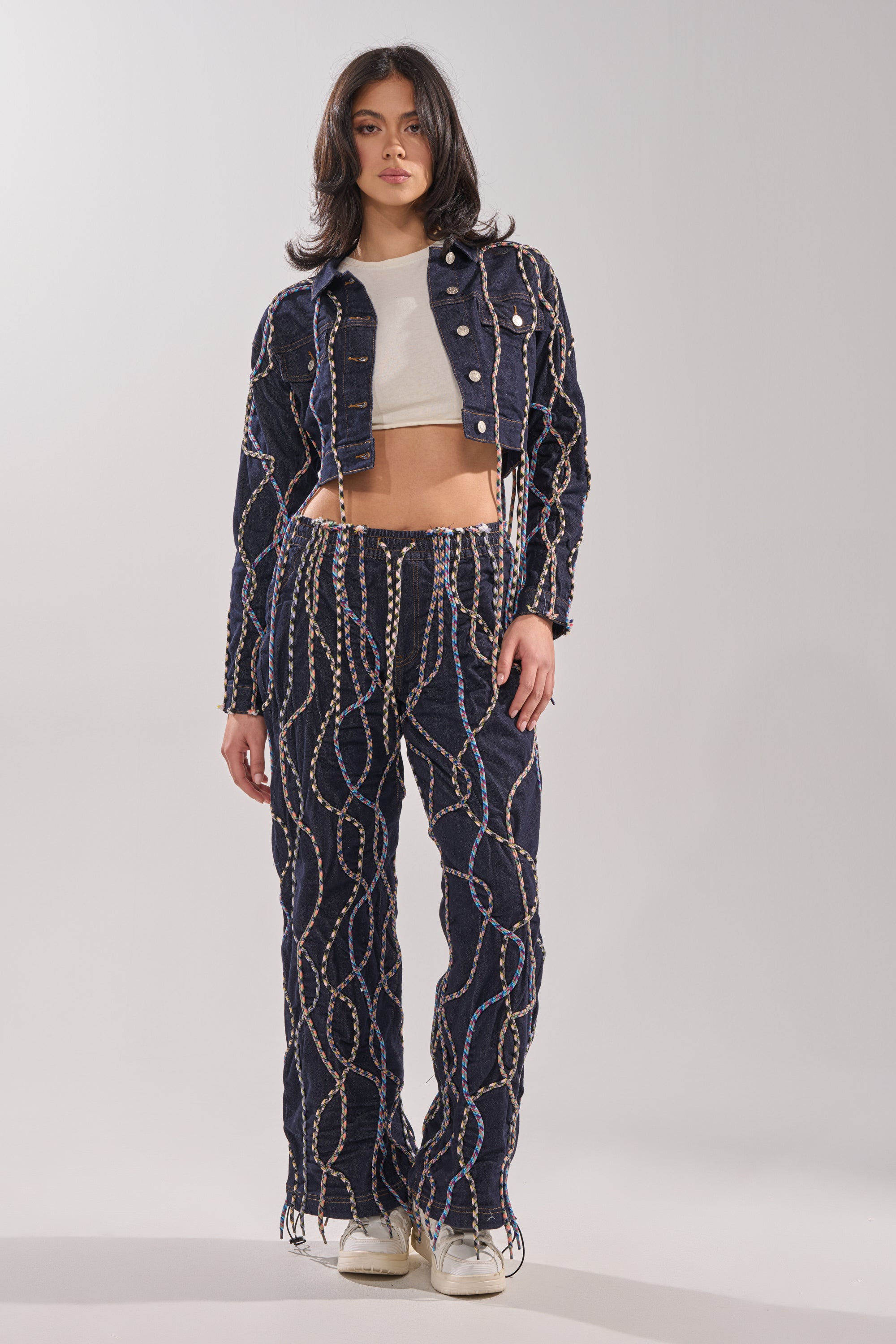 A woman with shoulder-length dark hair faces forward, wearing a white crop top, white sneakers, a cropped denim jacket, and CRAZY FOR YOU DENIM PANTS featuring a wavy chain-like pattern. 5