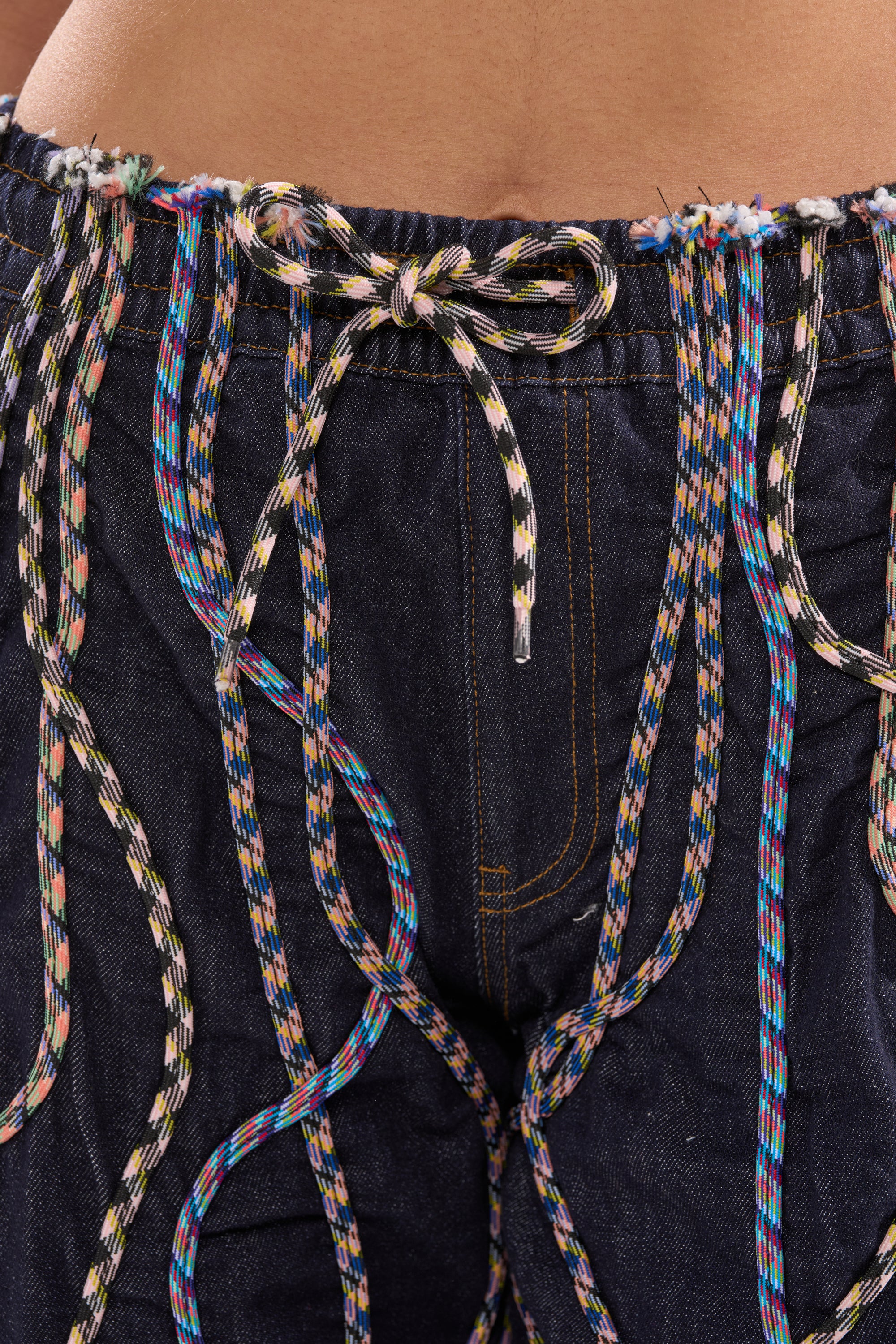 Close-up of the CRAZY FOR YOU DENIM PANTS: high-rise dark denim featuring colorful braided cords at the waist, multi-hued stitching, and vibrant rope detailing draped across the embellished design. 6