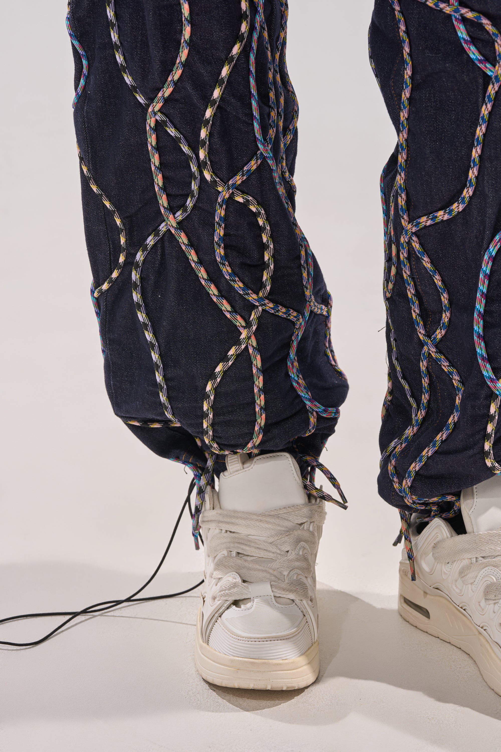 Close-up of a person wearing white sneakers and CRAZY FOR YOU DENIM PANTS, dark blue high-rise jeans embellished with multicolor rope loop patterns and slightly gathered at the ankles. 7