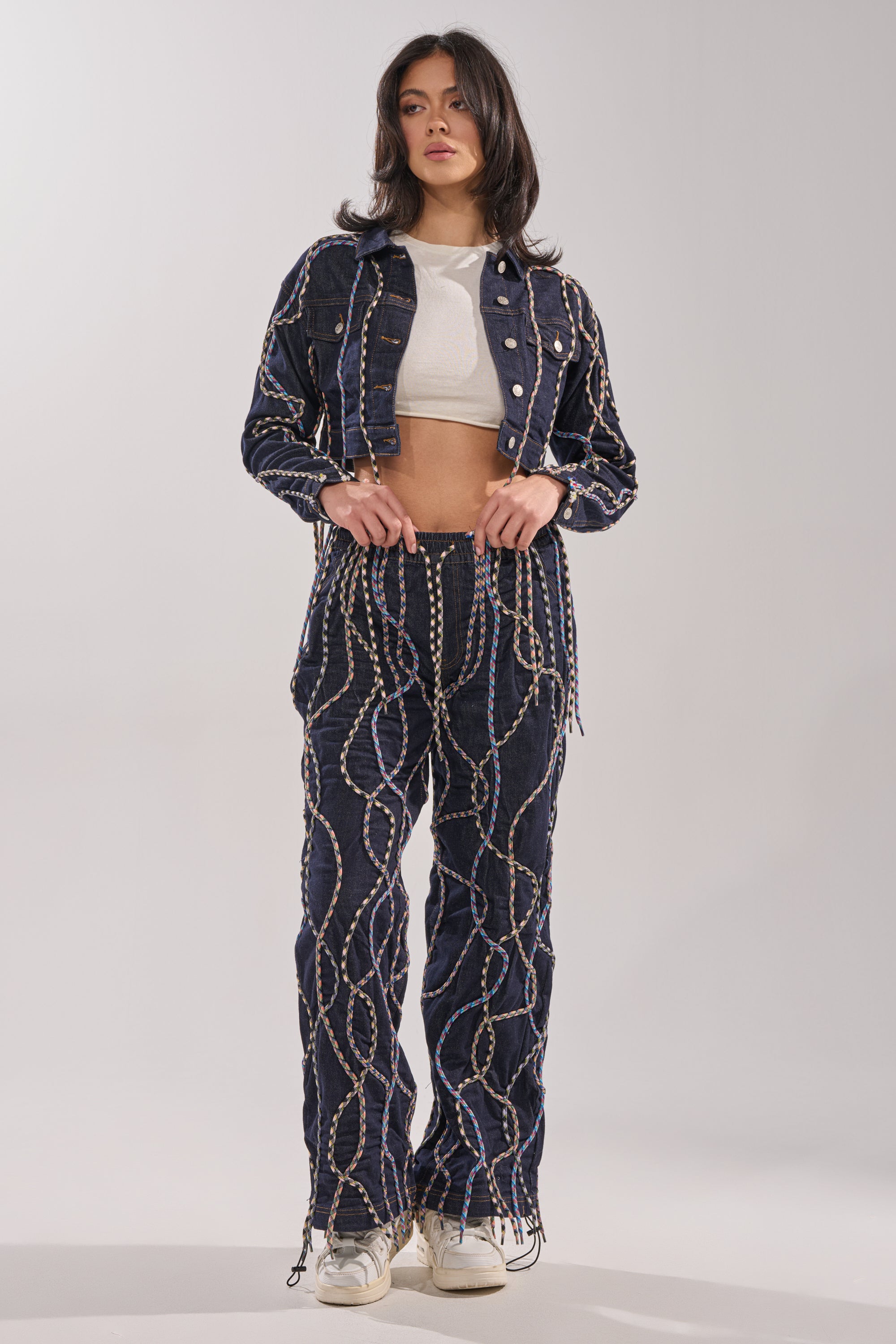 A woman faces forward, showcasing street style in the CRAZY FOR YOU DENIM JACKET with multicolor rope details and matching pants, paired with a white crop top and sneakers against a plain background. 0