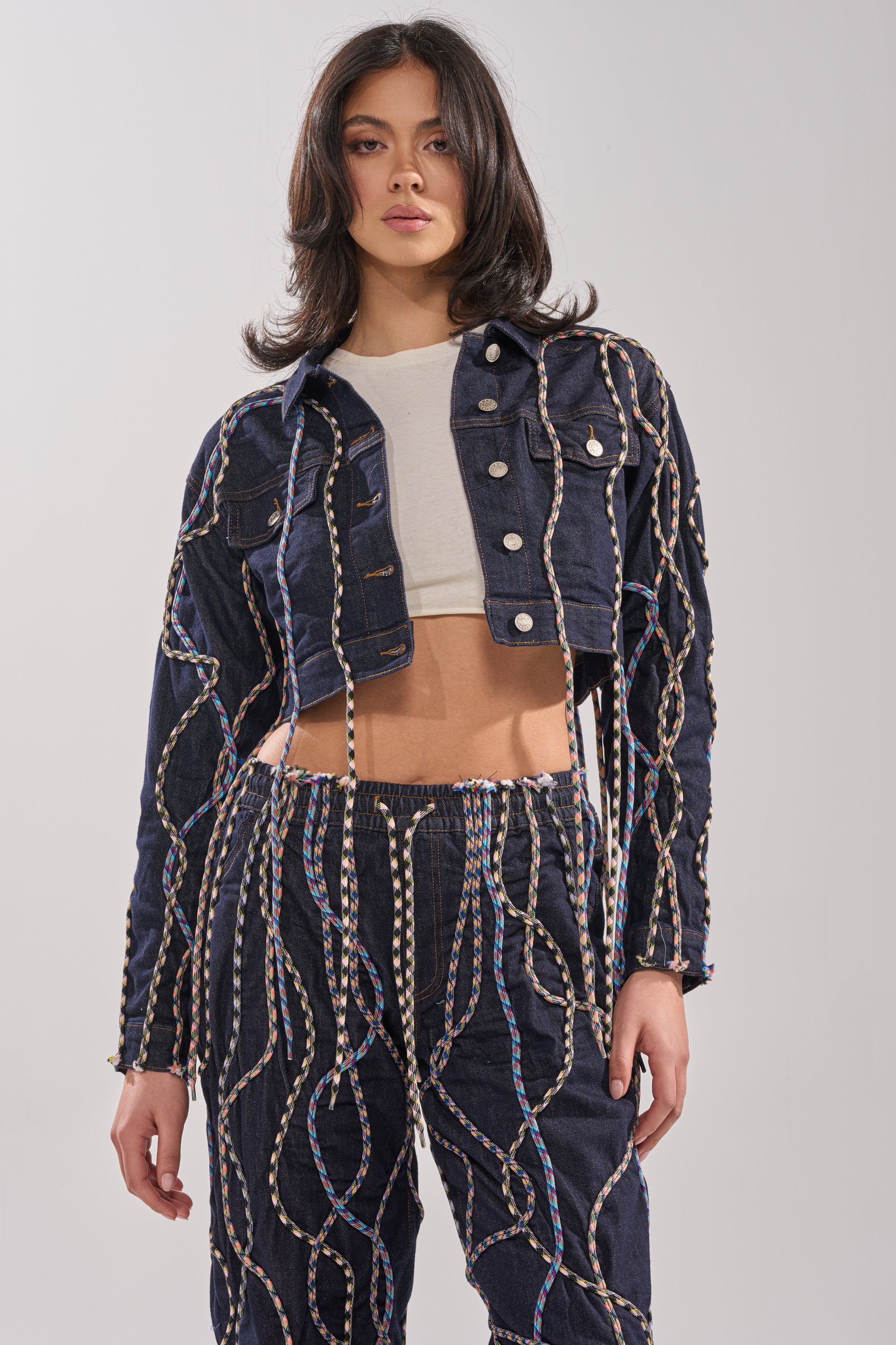 A woman wears the CRAZY FOR YOU DENIM JACKET, a cropped denim piece with colorful rope detailing in wavy patterns, paired with matching high-waisted pants for a bold streetstyle look against a plain light background. 1