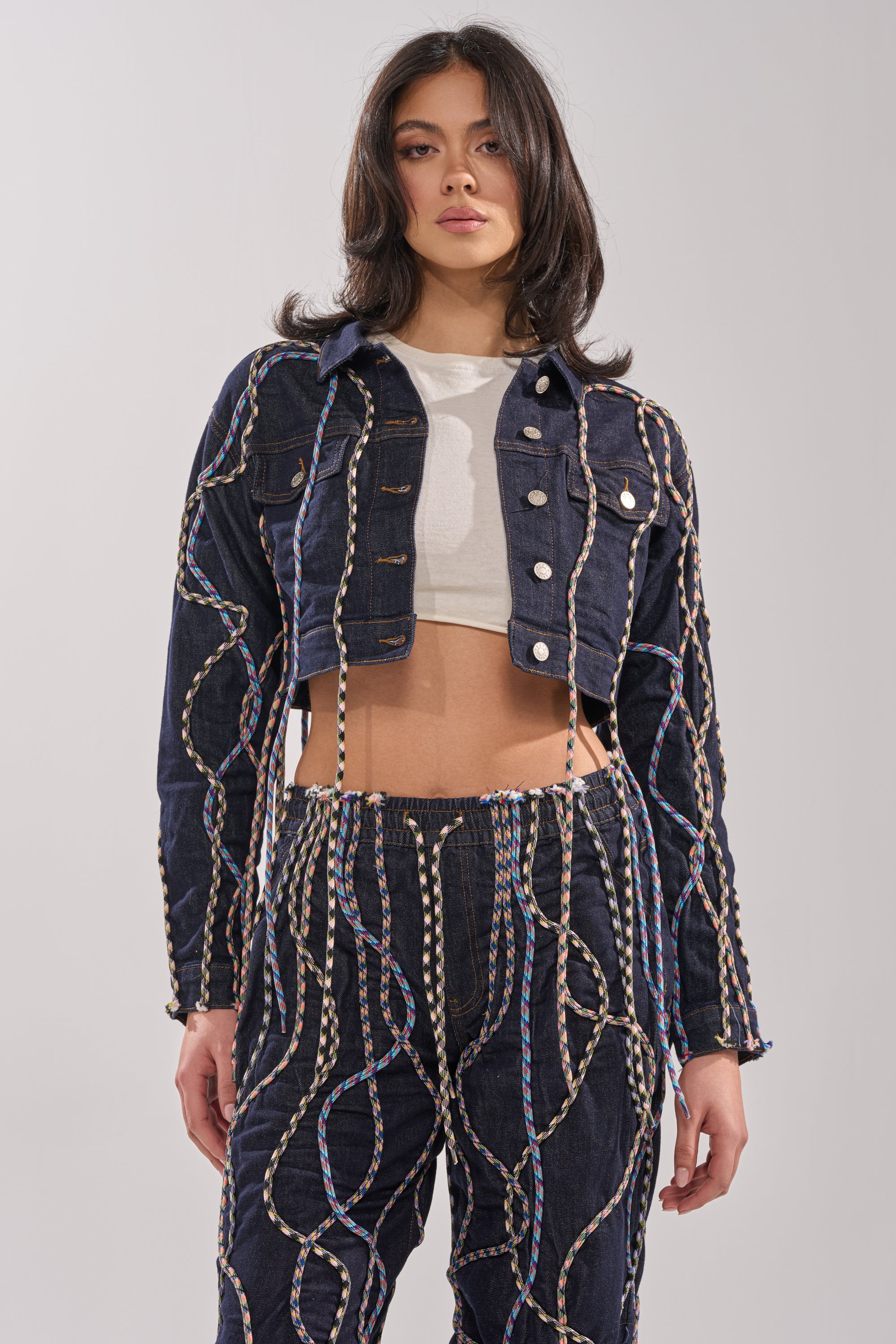 Wearing the CRAZY FOR YOU DENIM JACKET with matching high-waisted pants featuring rope details in blue, white, and tan over a white crop top, she embodies streetstyle flair against a plain light background. 2