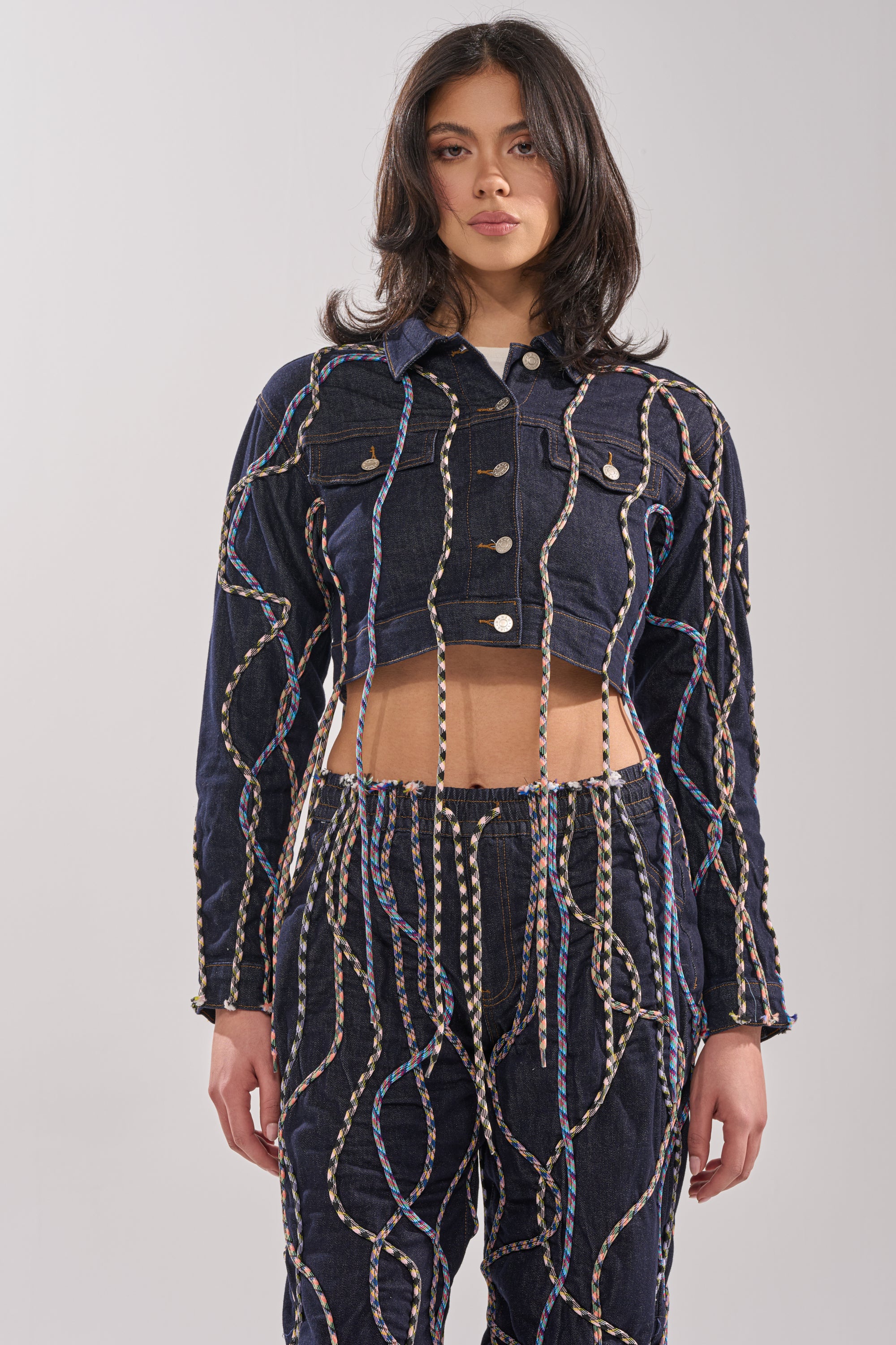 A woman with shoulder-length dark hair and a neutral expression styles the CRAZY FOR YOU DENIM JACKET, featuring colorful rope detailing, as part of a matching streetstyle set. 3