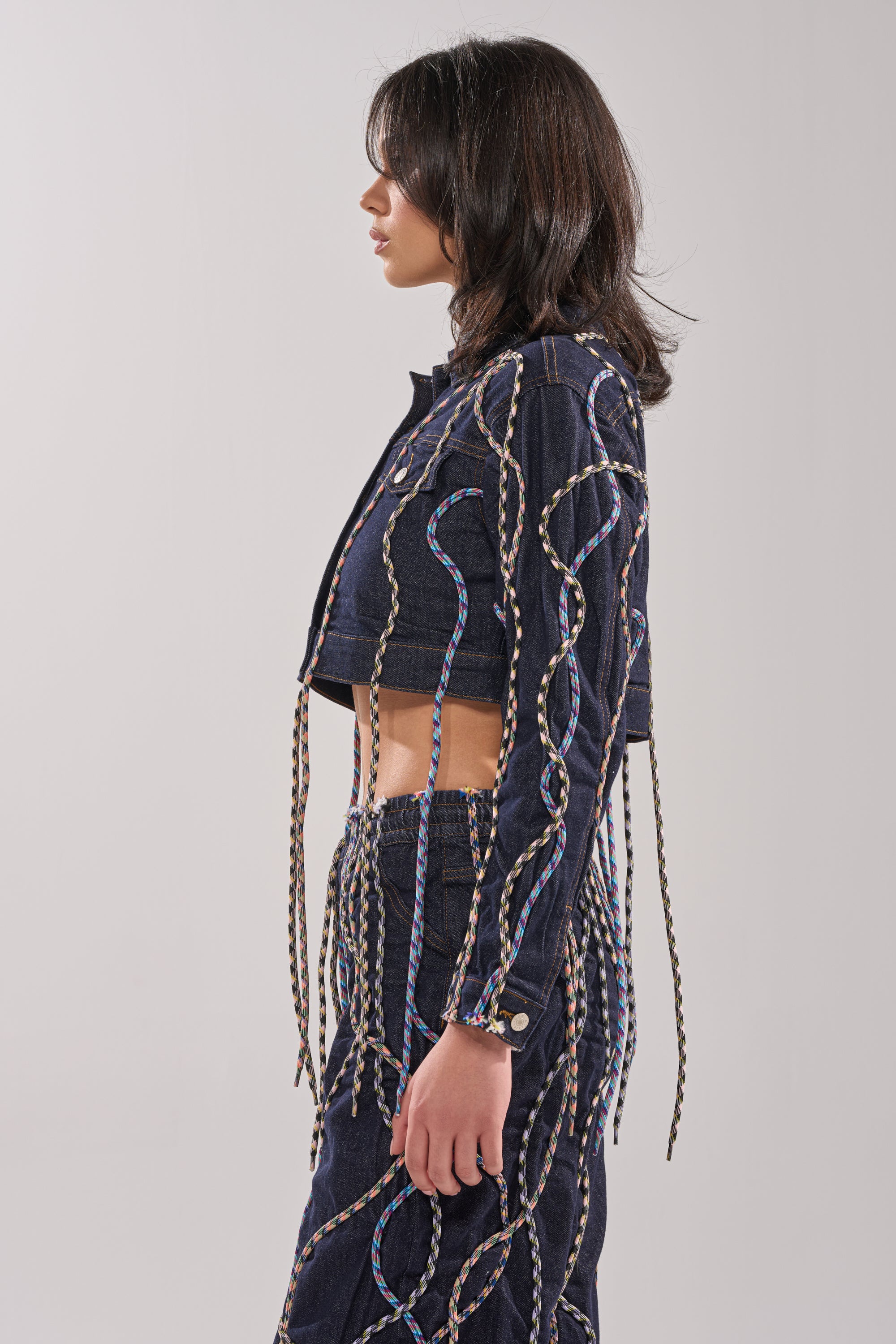 A woman in profile wears the CRAZY FOR YOU DENIM JACKET and matching bottoms, both featuring multicolor rope detailing in wavy patterns against a plain light gray background. 4
