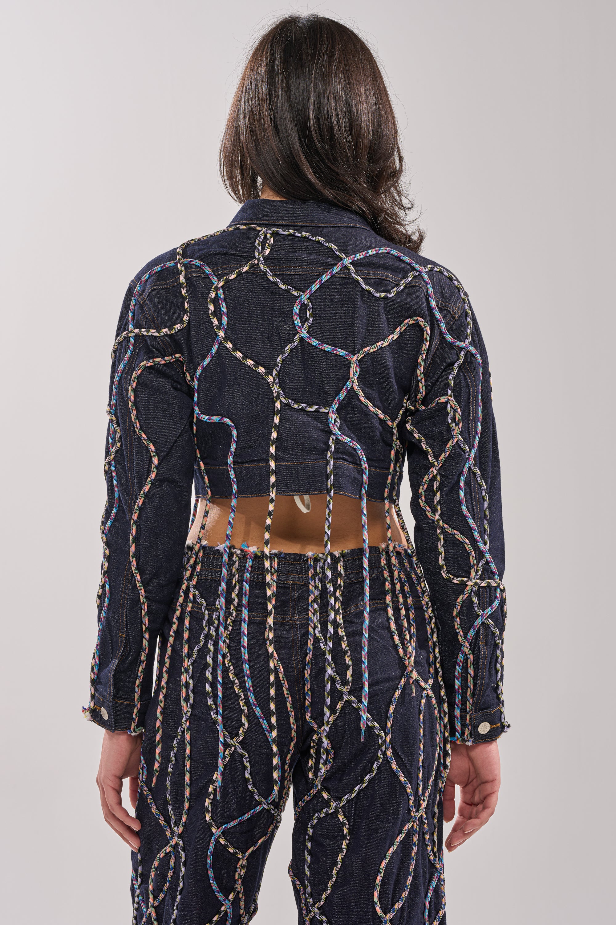 A person with shoulder-length dark hair is seen from behind wearing the CRAZY FOR YOU DENIM JACKET and pants featuring vibrant multicolor rope details, creating a standout streetstyle look against a plain, light background. 5
