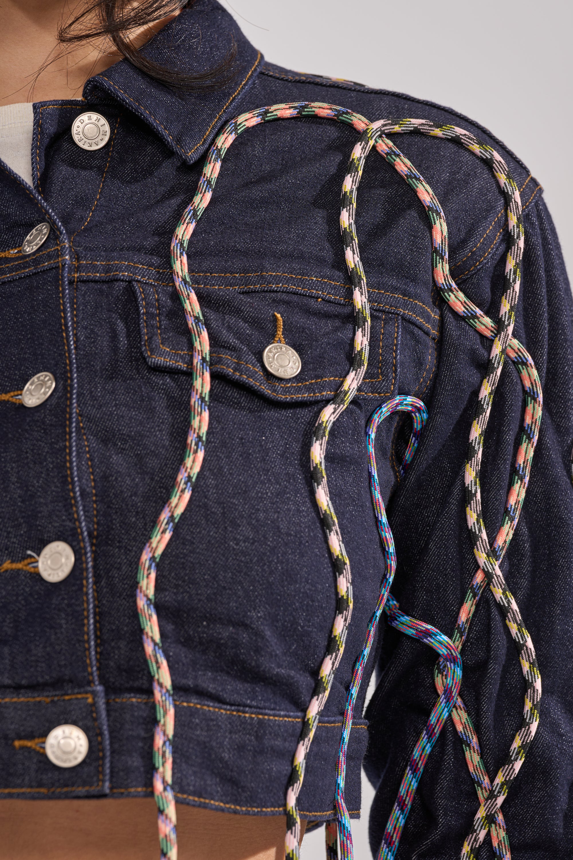 Close-up of the CRAZY FOR YOU DENIM JACKET featuring large silver buttons and multicolor rope detailing draped across the shoulder and front, giving a streetstyle edge to its dark blue cropped denim design. 7