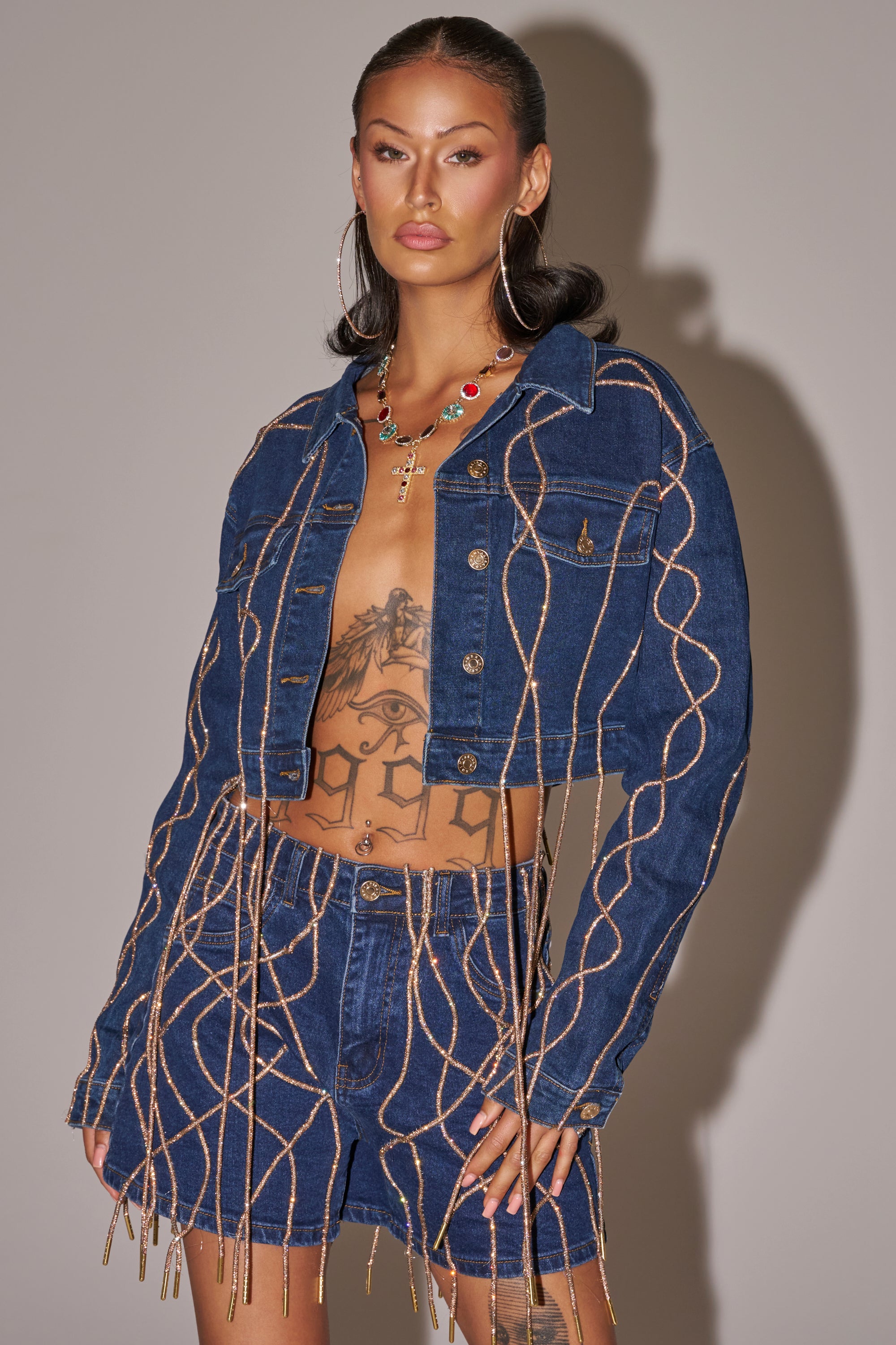 A woman with tattoos wears the NINA EMBELLISHED DENIM JACKET and matching shorts, featuring rhinestone details and gold chains. She accessorizes with hoop earrings, a jeweled necklace, and has sleek, shoulder-length dark hair. 1