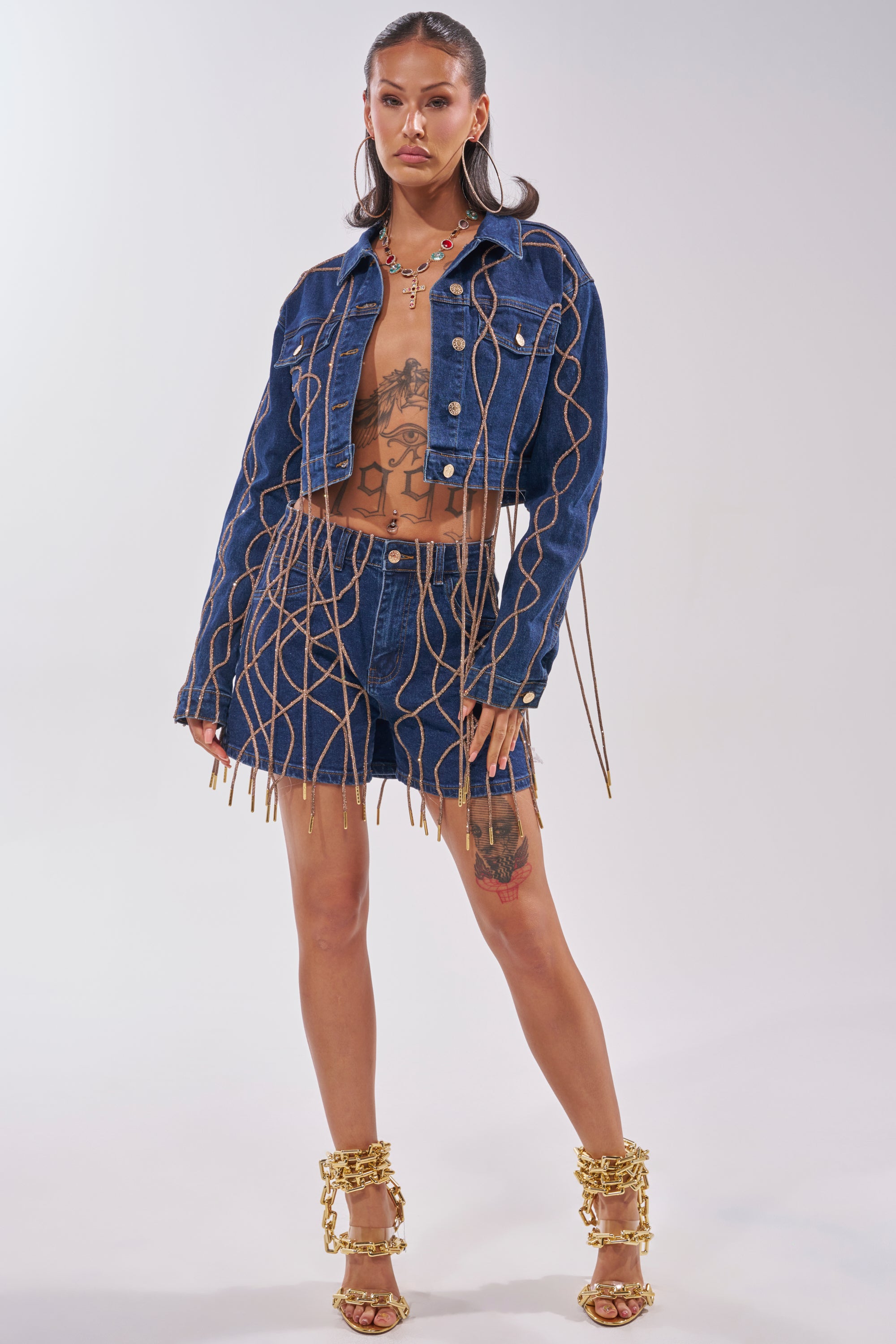 A woman poses in the NINA EMBELLISHED DENIM JACKET, featuring gold chain details. She accessorizes with large hoop earrings, a gold necklace, and gold chain-embellished heels. Her tattoos stand out against a plain white background. 5