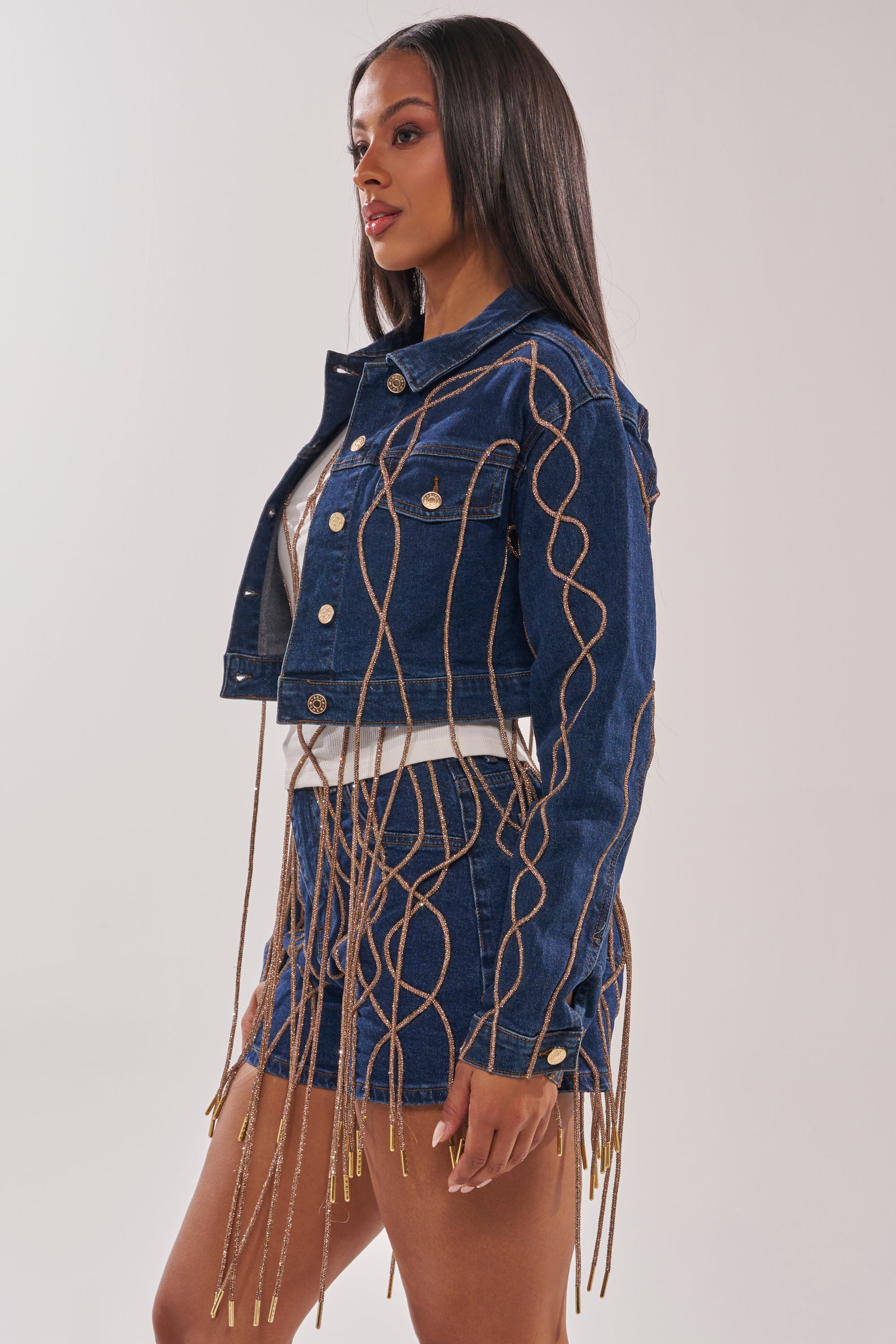 A woman stands against a plain light background wearing the NINA EMBELLISHED DENIM JACKET and matching shorts, both detailed with wavy gold fringe on the sleeves and hem. 2