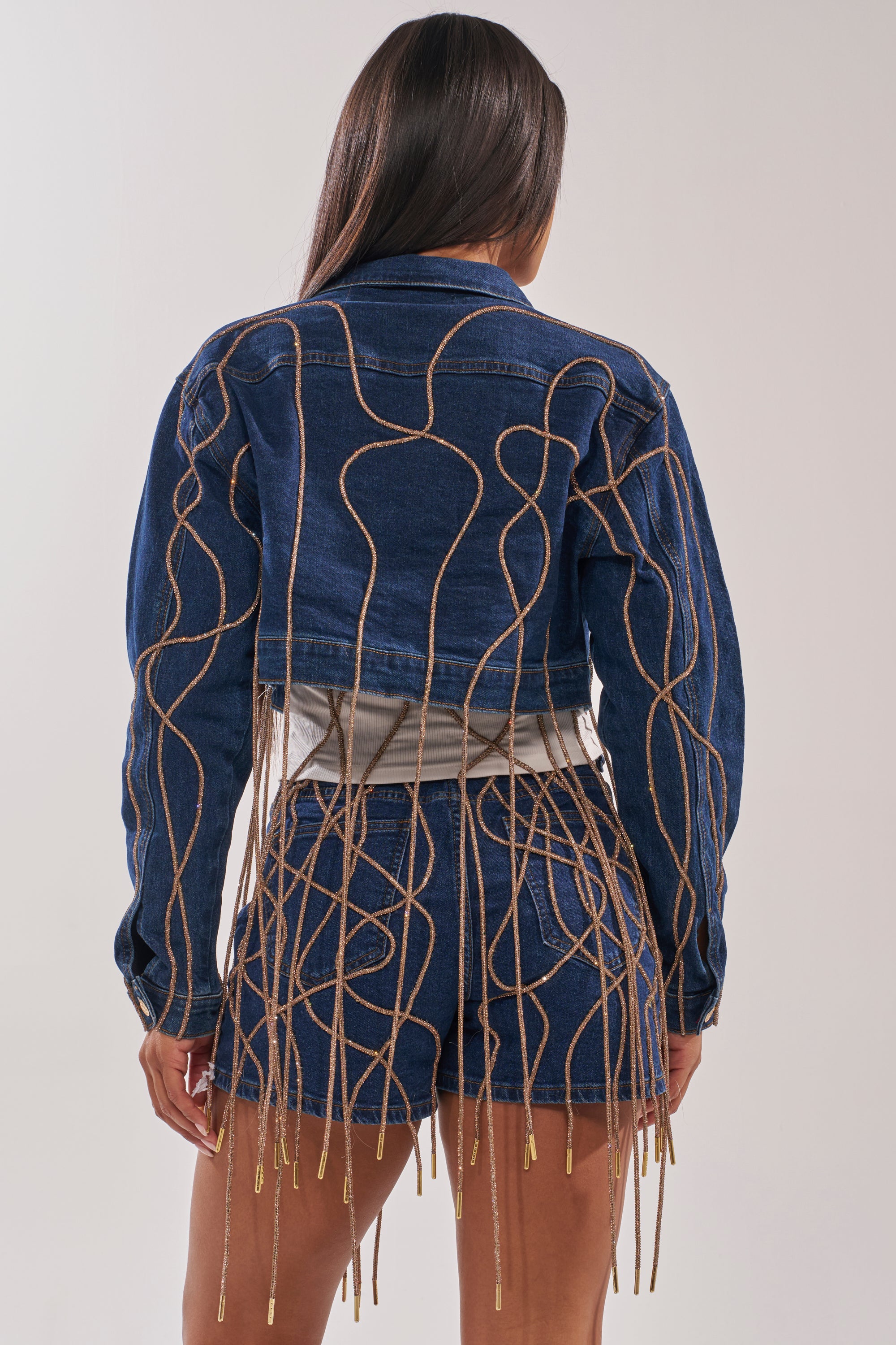 A person is seen from behind wearing the NINA EMBELLISHED DENIM JACKET and matching shorts, both in dark-wash denim with tan cords forming abstract patterns, highlighting the set’s detailed design. 3