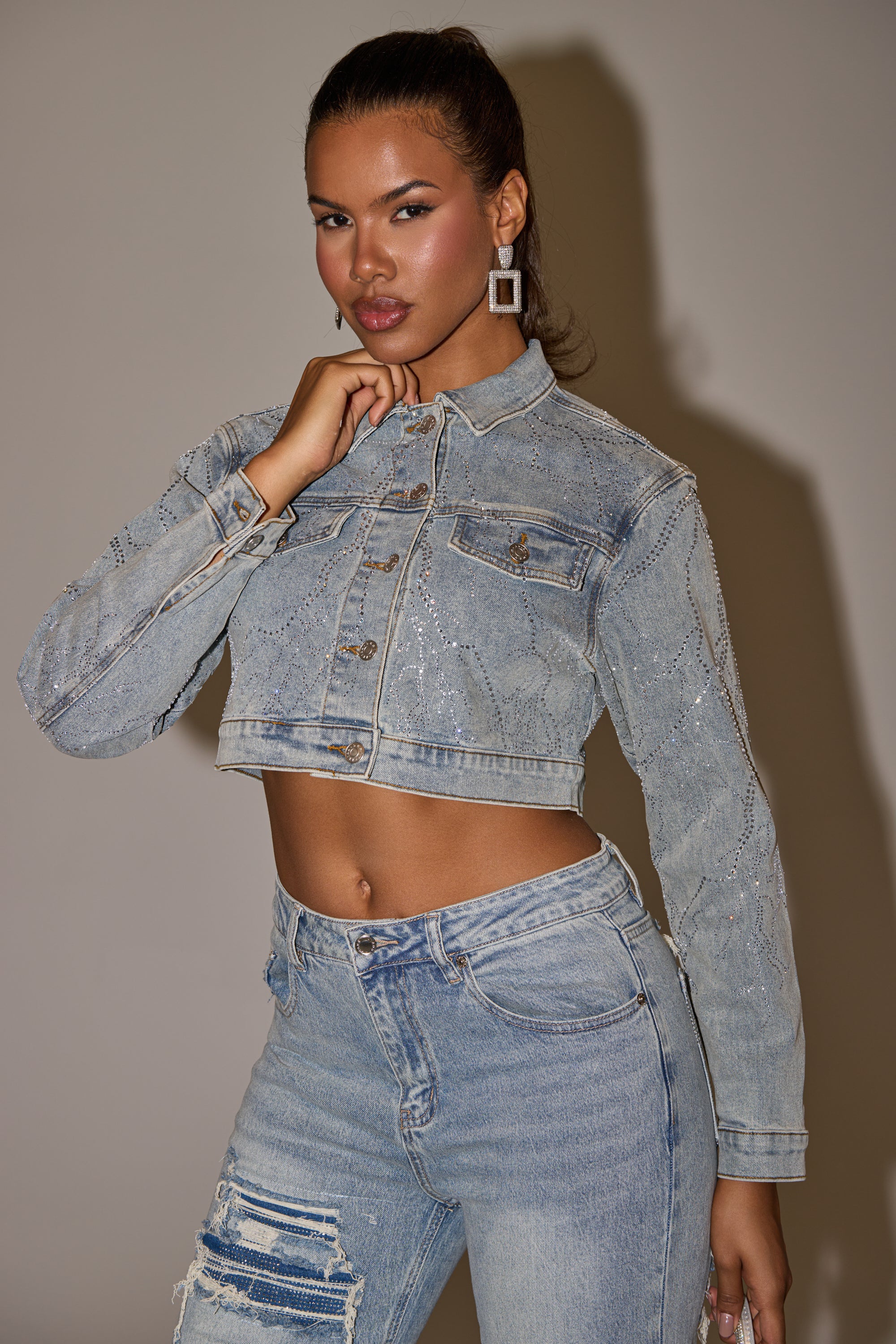 A woman with a confident expression and ponytail poses against a plain background wearing the DAZED RHINESTONE DENIM JACKET, featuring sparkly embellishments and paired with matching high-waisted, ripped jeans. 13