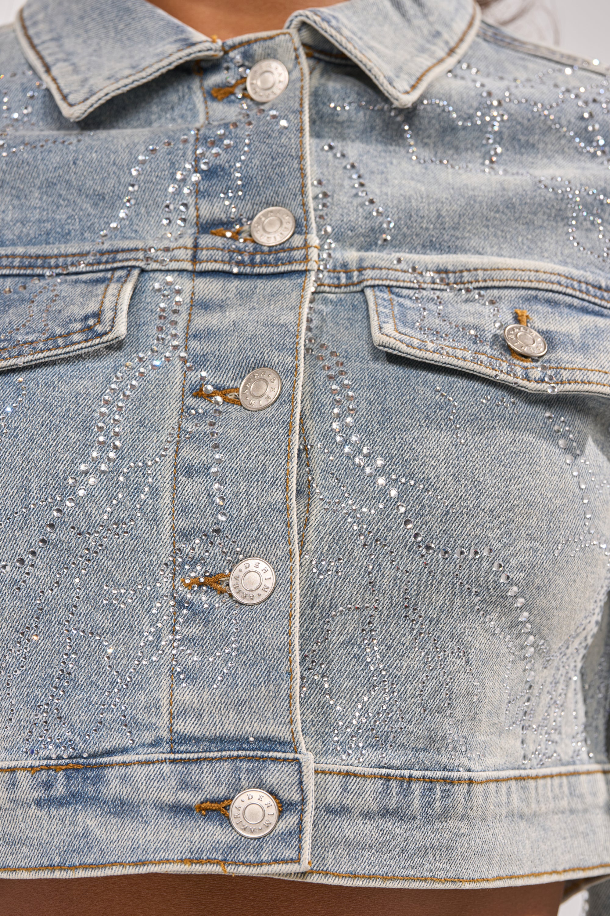 Close-up of the DAZED RHINESTONE DENIM JACKET in light blue, featuring metallic buttons, chest pockets, and scattered silver rhinestones in subtle wavy patterns. The jacket is worn open at the bottom, revealing bare skin. 14
