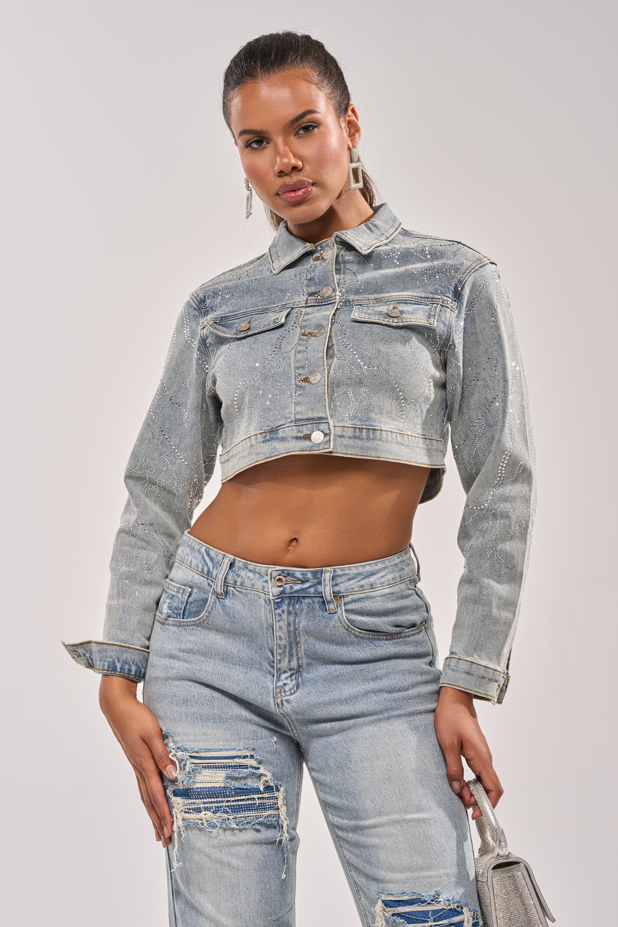 A woman poses confidently against a plain background in the DAZED RHINESTONE DENIM JACKET and matching distressed jeans, holding a small silver handbag. 7
