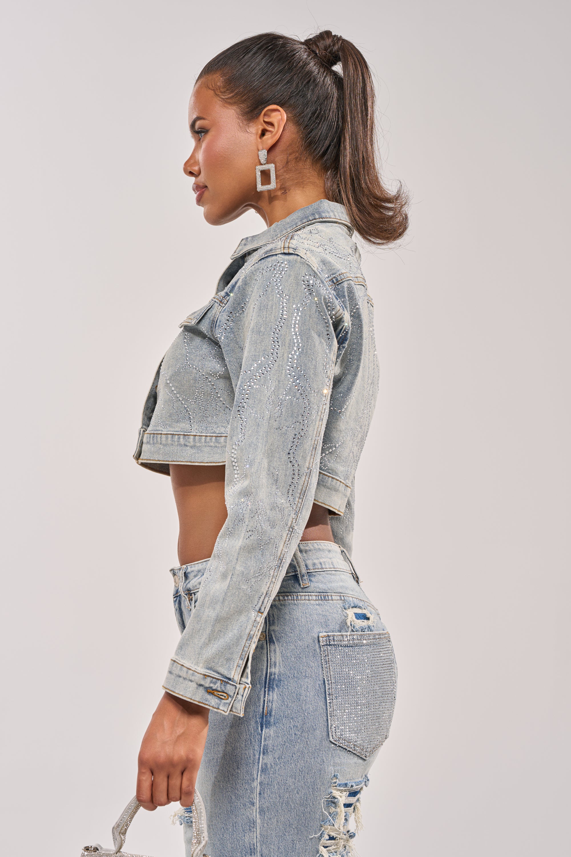 A woman in profile wears the DAZED RHINESTONE DENIM JACKET with matching ripped jeans. Her hair is styled in a high ponytail, and she holds a small bag, standing against a plain light gray background. 9