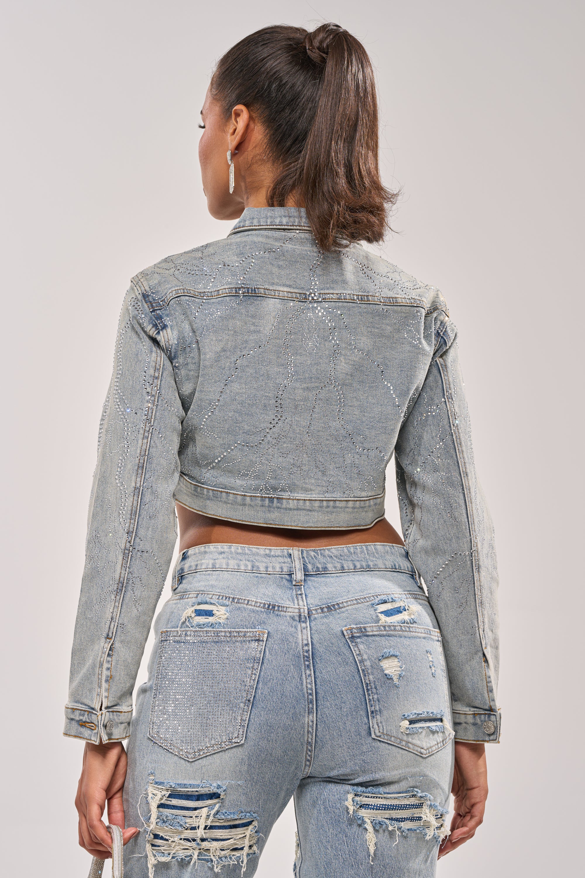 A woman with a high ponytail stands with her back to the camera, wearing the DAZED RHINESTONE DENIM JACKET and distressed light blue jeans, holding a small item in her left hand. 10