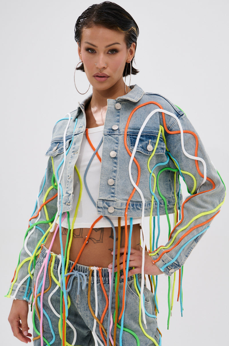 A woman in the WALK IT TALK IT DENIM JACKET, featuring 3D rope details, pairs it with jeans with colorful accents and fringe. She stands against a white background, wearing large hoop earrings and sporting slicked-back hair. 0