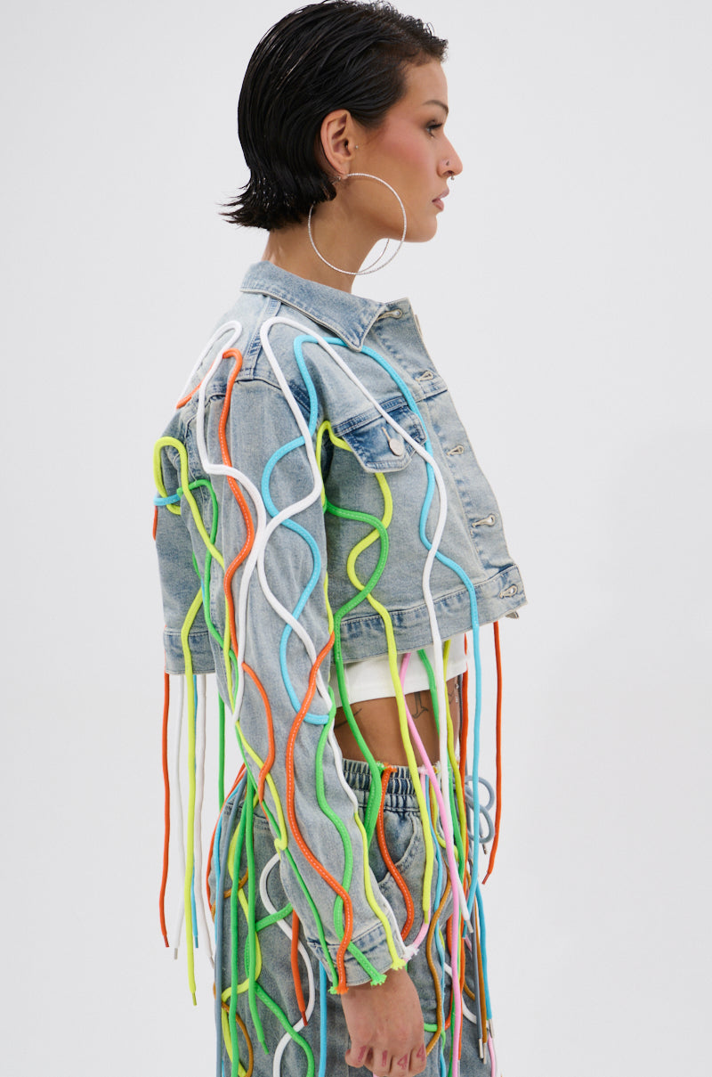 A woman with short dark hair wears the WALK IT TALK IT DENIM JACKET and matching jeans, both featuring colorful 3D rope detailing. She stands in profile against a plain white background, wearing large hoop earrings. 1