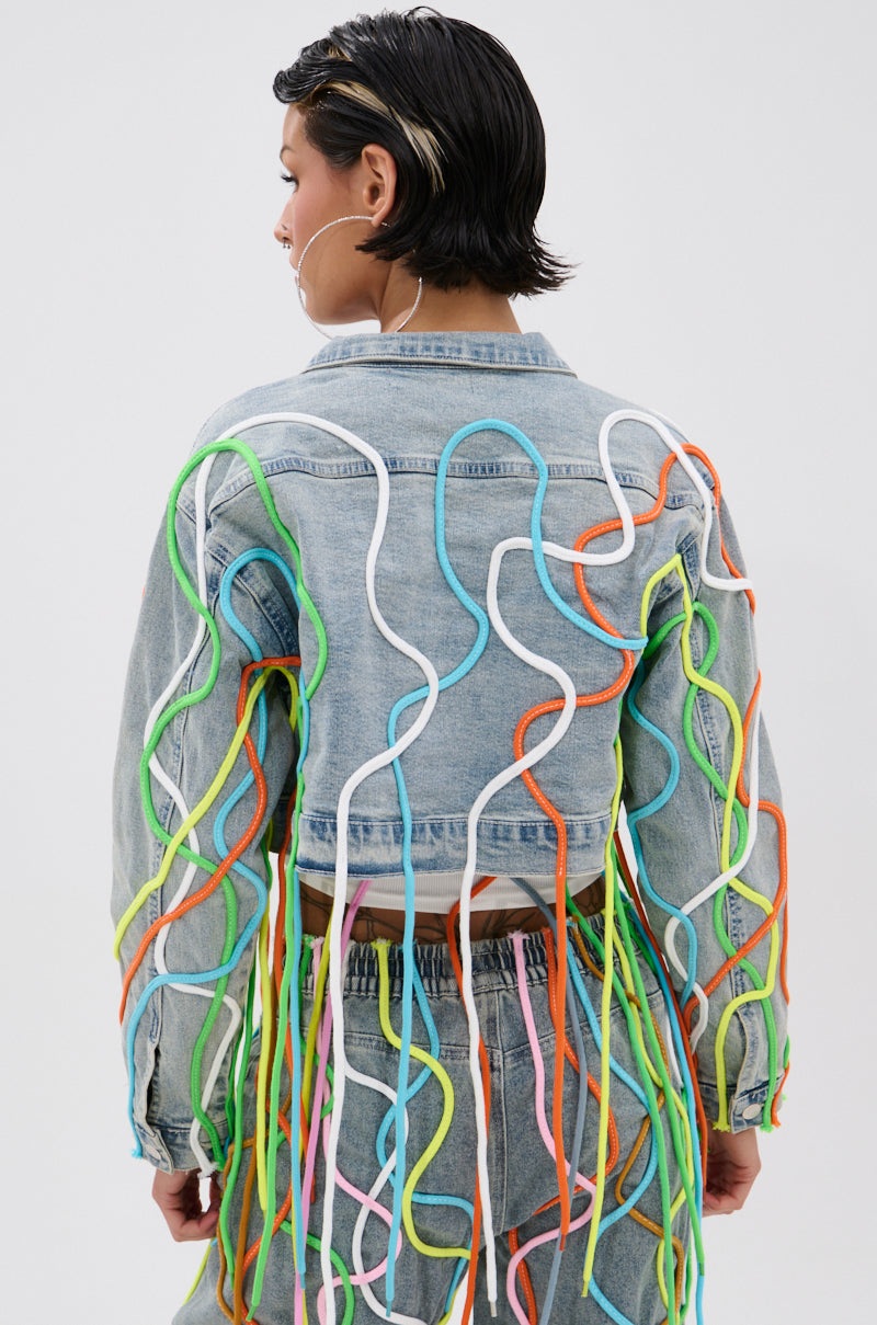 A person with short, dark, wet-look hair is seen from behind wearing the WALK IT TALK IT DENIM JACKET, which features 3D rope detailing—colorful wavy cords in green, orange, blue, yellow, and white on the shoulders and back. 2