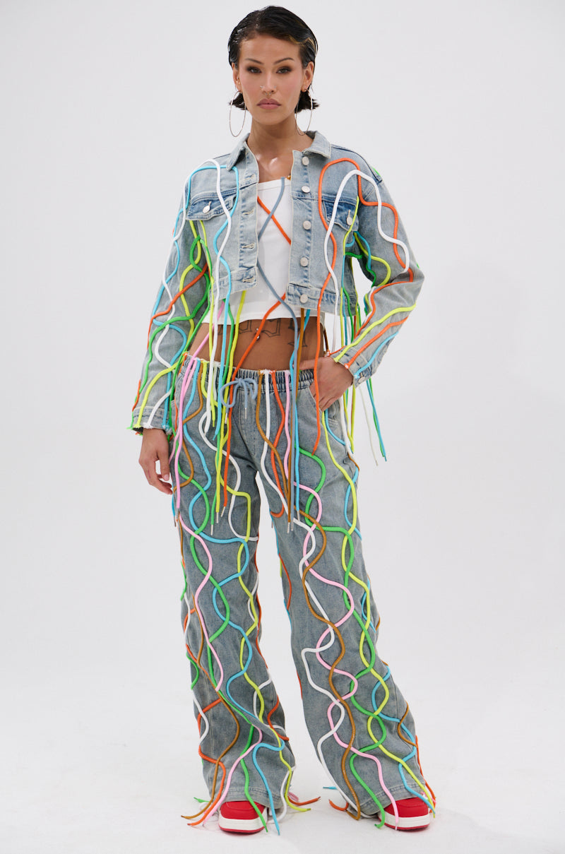 A woman stands confidently in the WALK IT TALK IT DENIM JACKET and matching jeans with colorful 3D rope details, a white crop top, and red sneakers. Her hair is slicked back as she poses against a plain background. 3