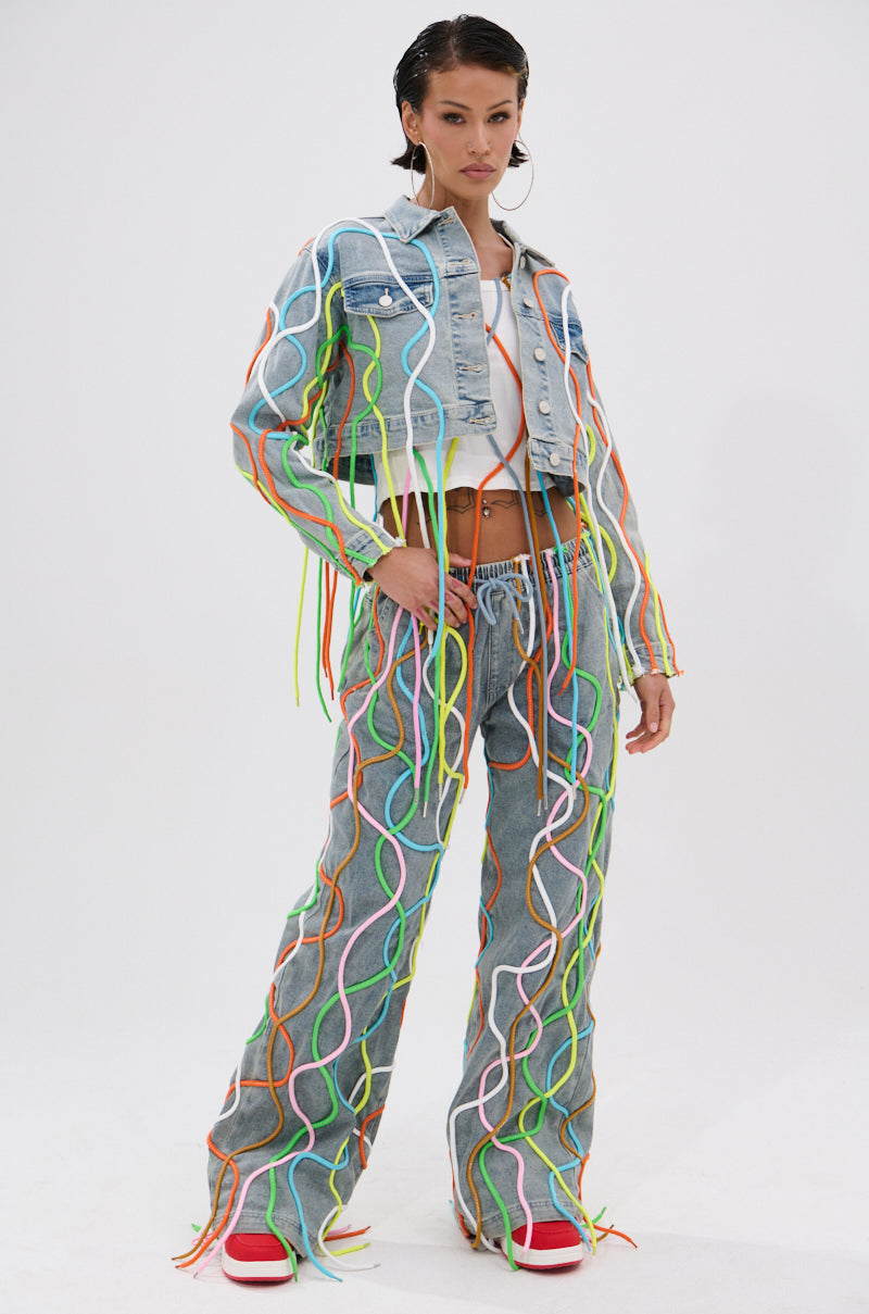 A woman models the WALK IT TALK IT DENIM JACKET with 3D colorful rope details, paired with matching jeans, a white crop top, red shoes, and silver hoop earrings against a plain white background. 4