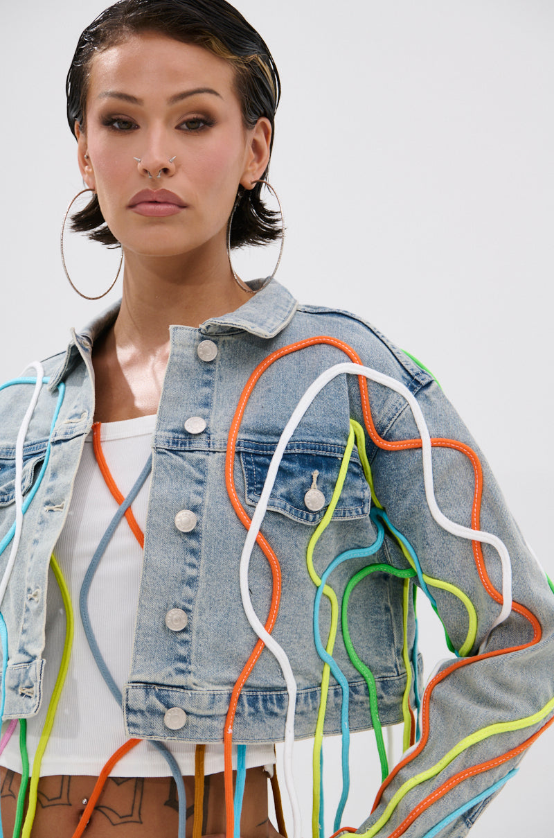 A person with short dark hair poses against a plain background in the WALK IT TALK IT DENIM JACKET, which features vibrant 3D rope detailing. They wear a white crop top, hoop earrings, a nose ring, and display torso tattoos. 5
