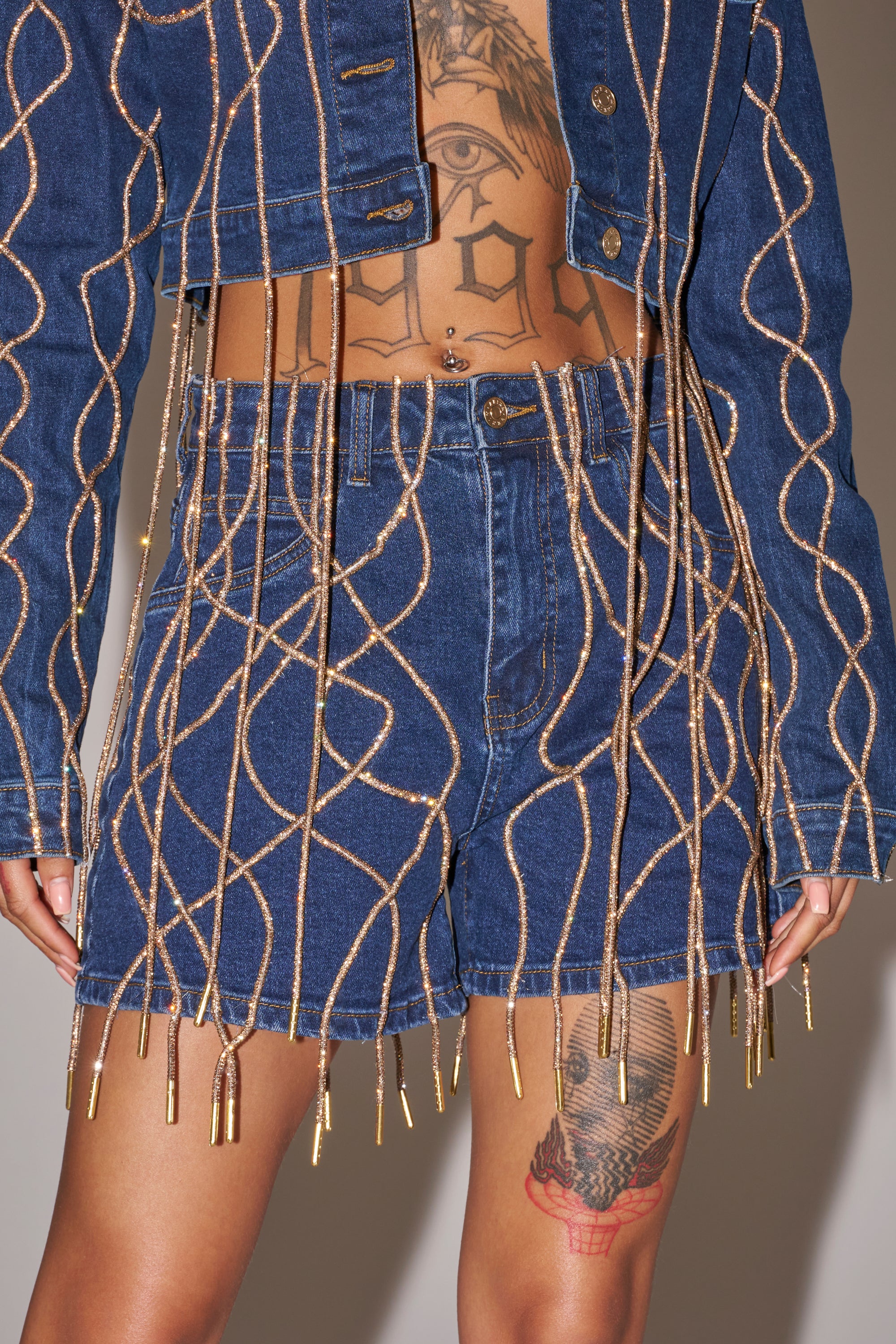 A person shows tattooed skin while wearing the NINA EMBELLISHED BERMUDA SHORT, featuring rhinestone accents and gold cord details. Photo is cropped to highlight the lower torso and upper legs. 0