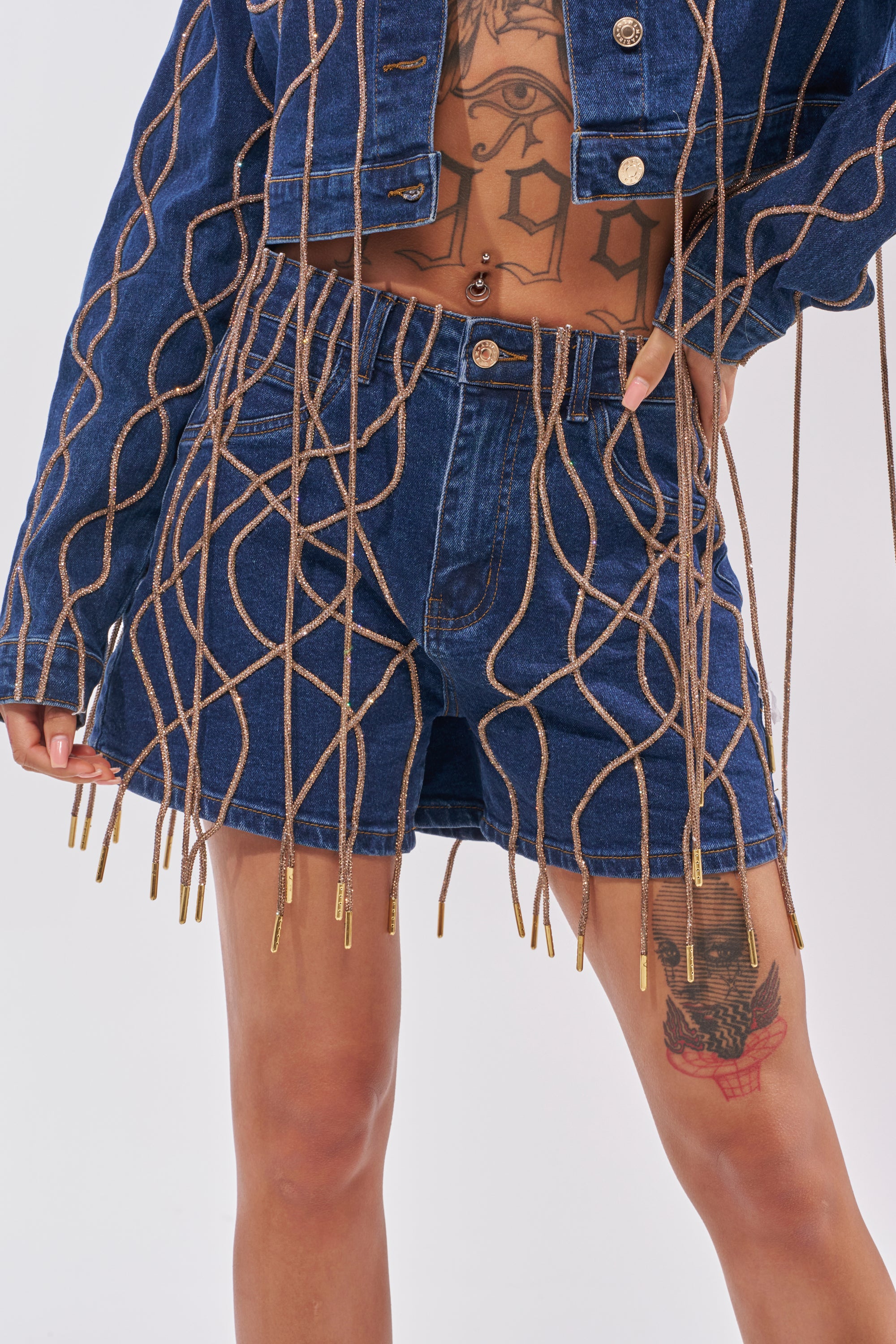Wearing a denim jacket and the NINA EMBELLISHED BERMUDA SHORT with tan rope-like fringe, this person shows off midriff and thigh tattoos. 8